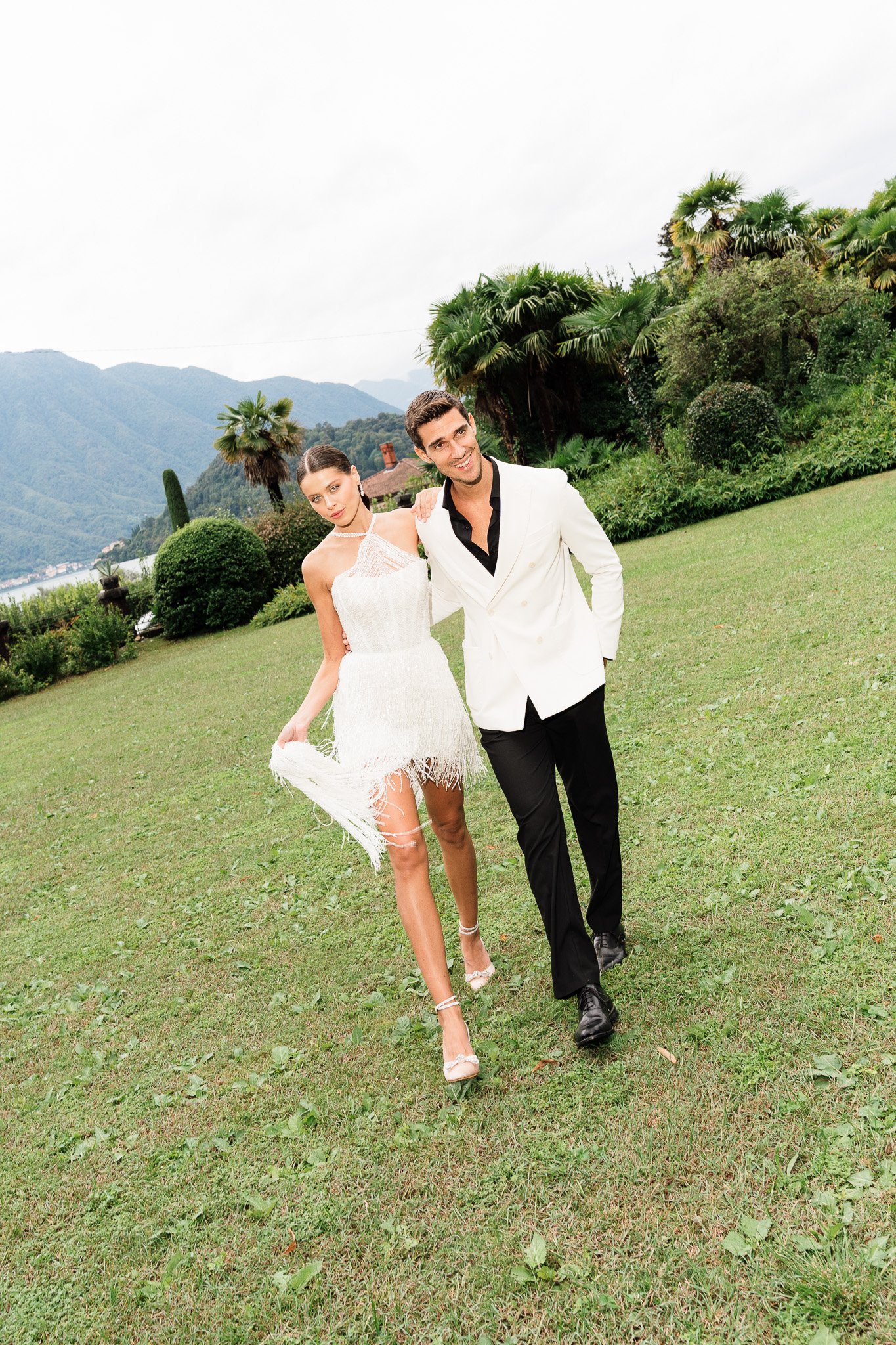 Ein Mann und eine Frau spazieren auf einer Wiese mit einem Rücken an das Wasser und Berge im Hintergrund. Die Frau trägt ein weißes, schulterfreies Kleid und hohe Schuhe, der Mann trägt einen weißen Anzug mit schwarzen Hosen.