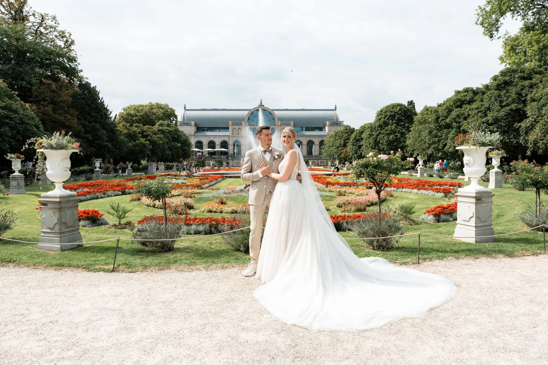 Ein Brautpaar in Hochzeitskleidung steht vor einem blumenreichen Garten und einem historischen Gebäude im Hintergrund.