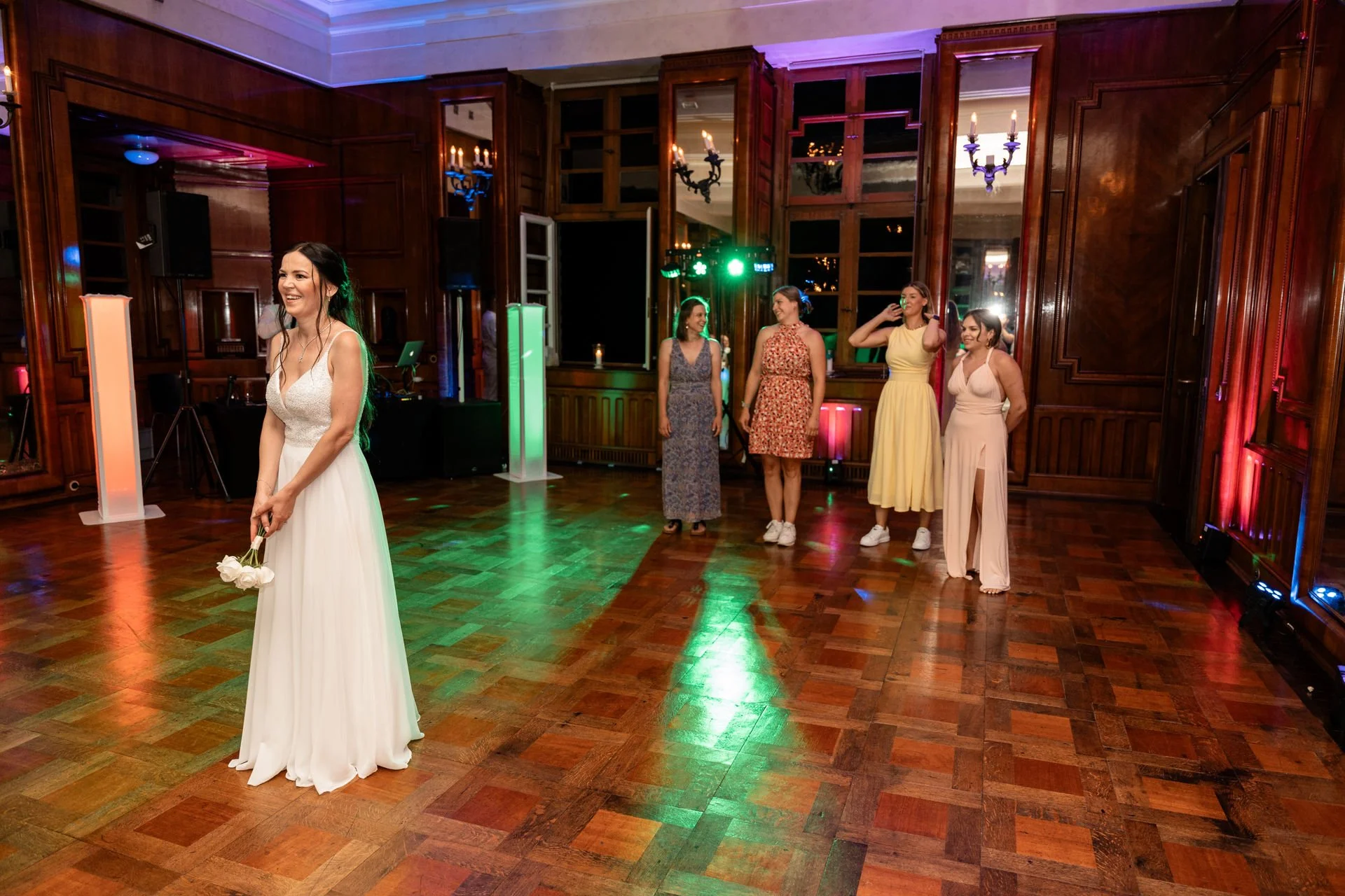 Eine Hochzeitsfeier mit einer Braut in einem weißen Kleid, die in einem festlich dekorierten Raum steht, während vier Frauen im Hintergrund lachen und sich unterhalten.