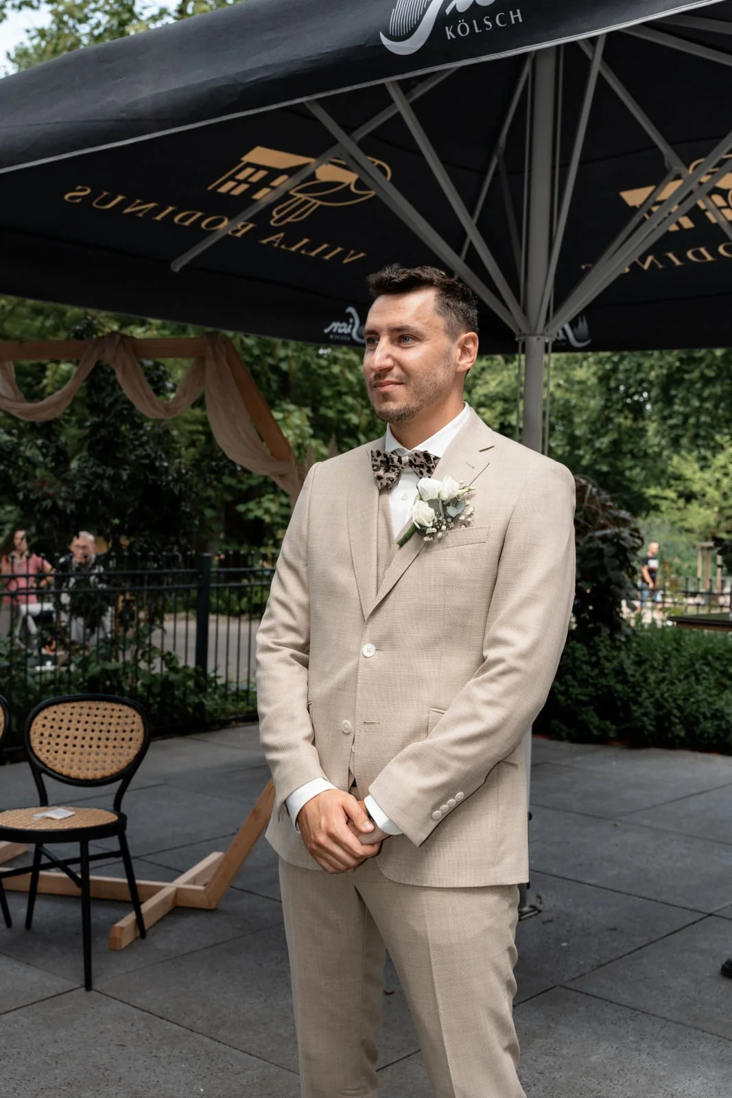 Ein Mann steht in einem beige Anzug mit einer Fliege und einem weißen Hemd bei einer Hochzeit im Freien, unter einem großen schwarzen Sonnenschirm mit Goldschrift.