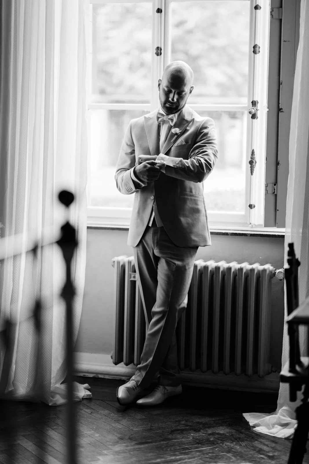 Ein Mann in Hochzeitsanzug steht vor einem Fenster, schaut auf seine Armbanduhr, im Raum sind Vorhänge und eine Heizkörper sichtbar.