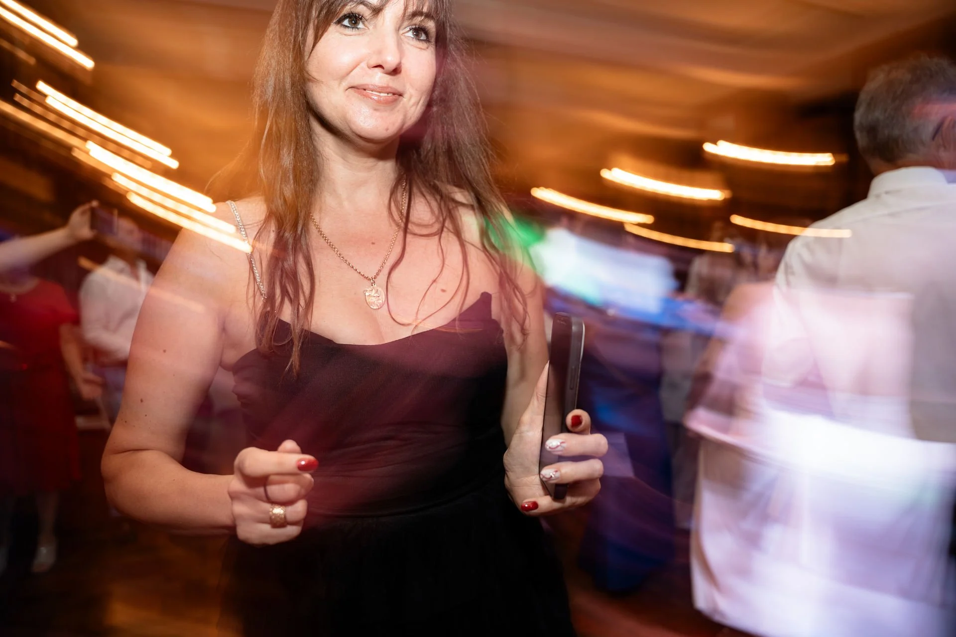 Frau in schwarzem Kleid hält ein Smartphone in der Hand bei einer Party, im Hintergrund bewegen sich andere Gäste bei Disco-Beleuchtung.
