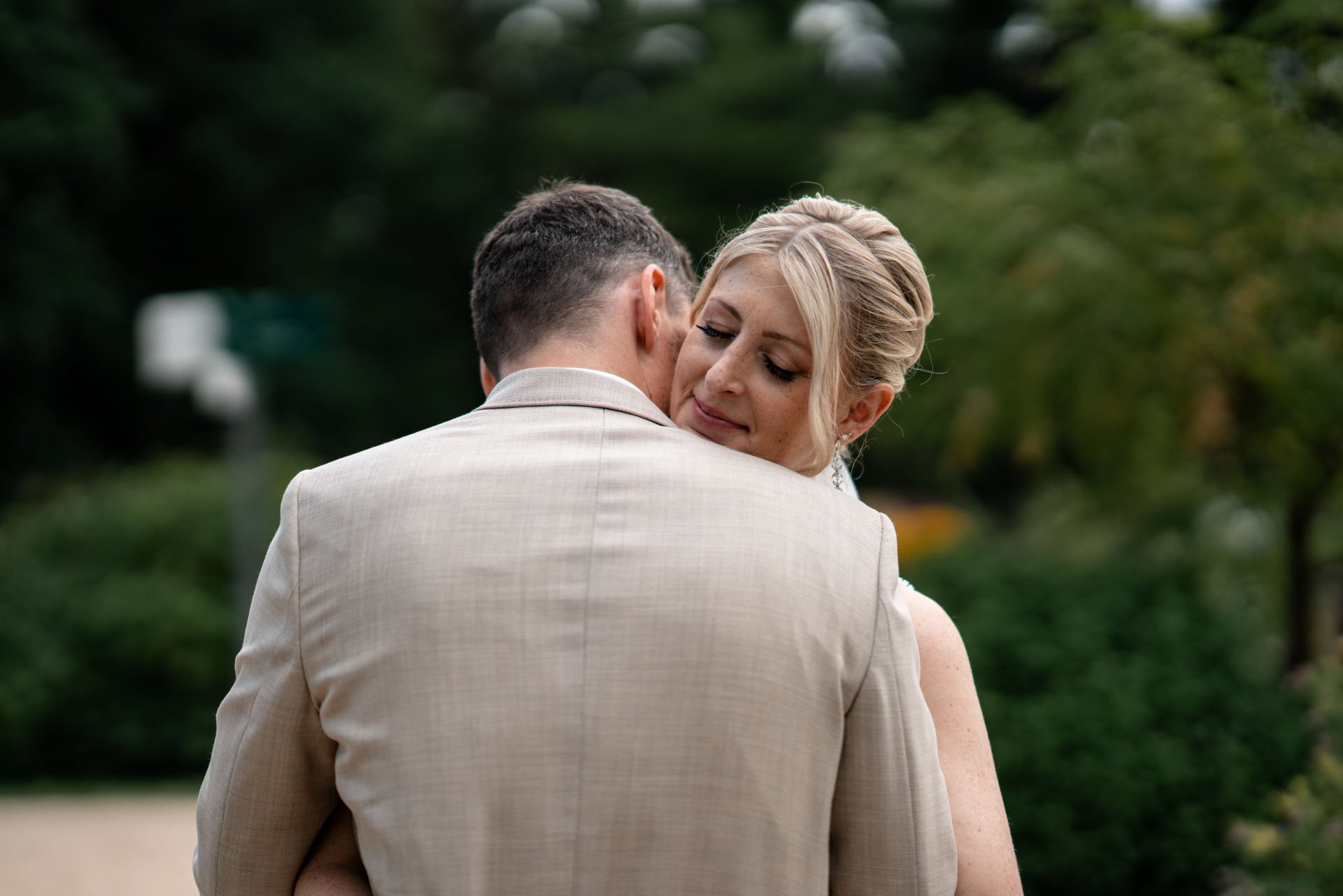 Ein Mann und eine Frau umarmen sich, vermutlich bei einer Hochzeit, im Freien mit grüner Baumkulisse im Hintergrund.