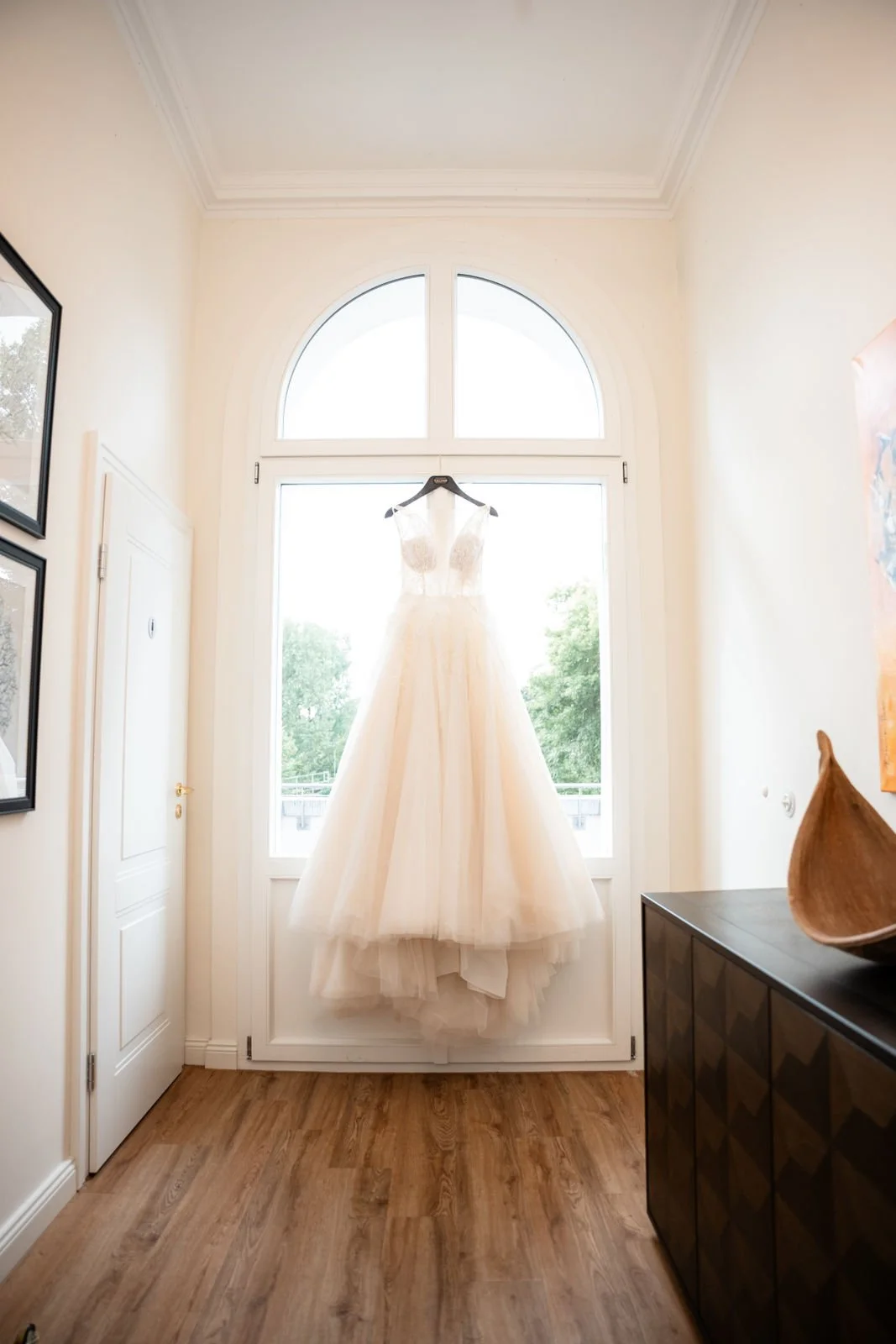 Brautkleid hängt vor einem großen Fenster in einem hellen Raum mit Holzfußboden.