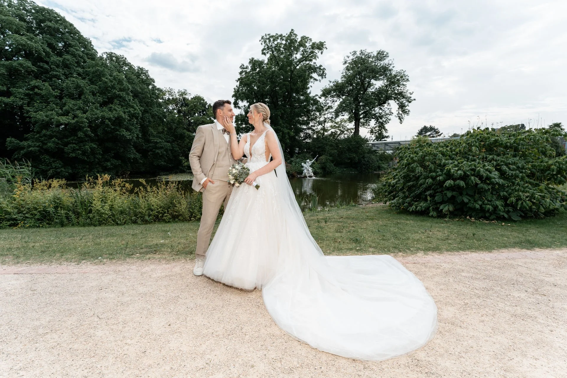 Ein Brautpaar steht am Ufer eines Sees, umgeben von Bäumen, bei einem Hochzeitsshooting. Sie trägt ein weißes Hochzeitskleid mit langem Schleier, er trägt einen beige Anzug, sie schaut sich liebevoll an.