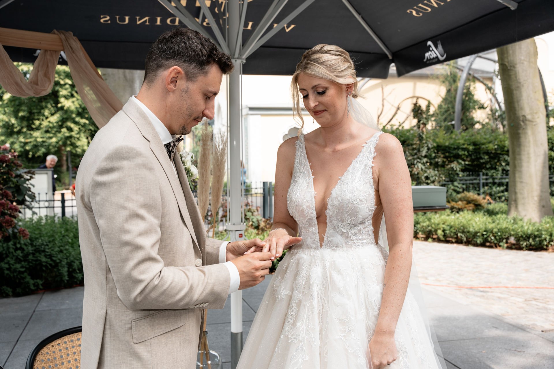 Ein Mann in einem hellen Anzug und eine Frau in einem weißen Hochzeitskleid bei einer Standesamtlichen Trauung im Freien, die sich gegenseitig das Ehering tauschen.