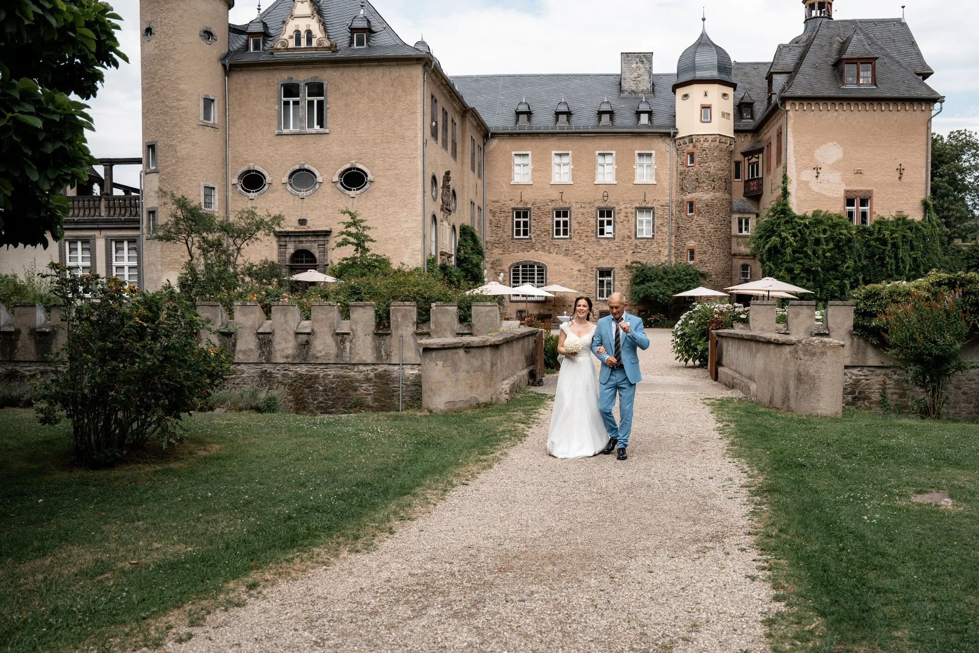 Ein Brautpaar spaziert vor einem historischen Schloss im Garten, die Braut trägt ein weißes Kleid und hält einen Blumenstrauß, der Bräutigam trägt einen blauen Anzug.