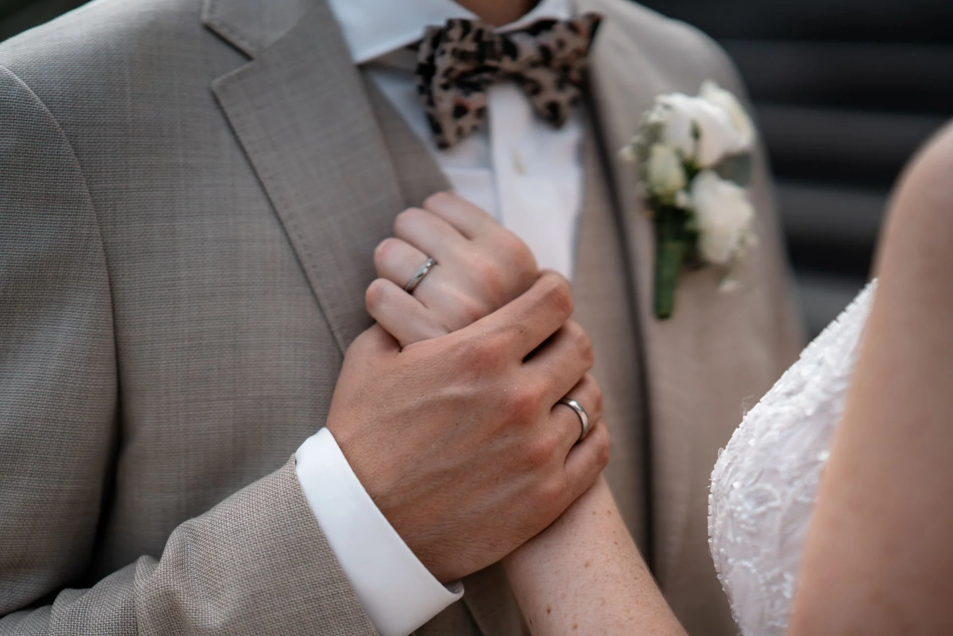 Nahaufnahme von einem Ehepaar bei der Hochzeit, das sich die Hände hält, mit Eheringen. Mann trägt Anzug mit einer Note an einem Oberteil, die Frau trägt ein weißes Kleid mit Stickerei.