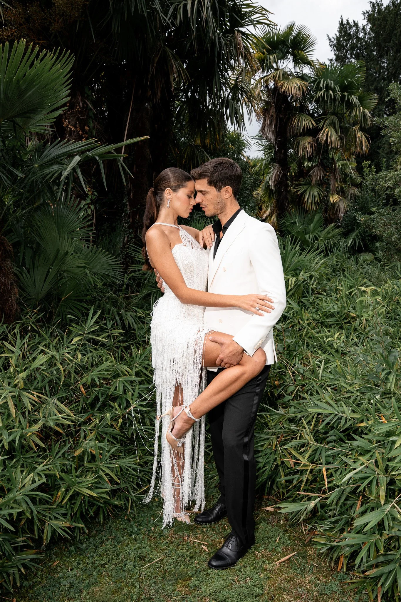 Ein Paar in formeller Kleidung steht sich inmitten von grünen Pflanzen und Bäumen gegenüber, die Partner berühren sich. Das Mädchen trägt ein weißes, schulterfreies Kleid mit Fransen, das man kaum im Blick hat, weil sie darin die Beine lindert und die Arme in die Ehe des Partners legt. Der junge Mann trägt einen weißen Anzug mit schwarzen Hosen und einem schwarzen Hemd.