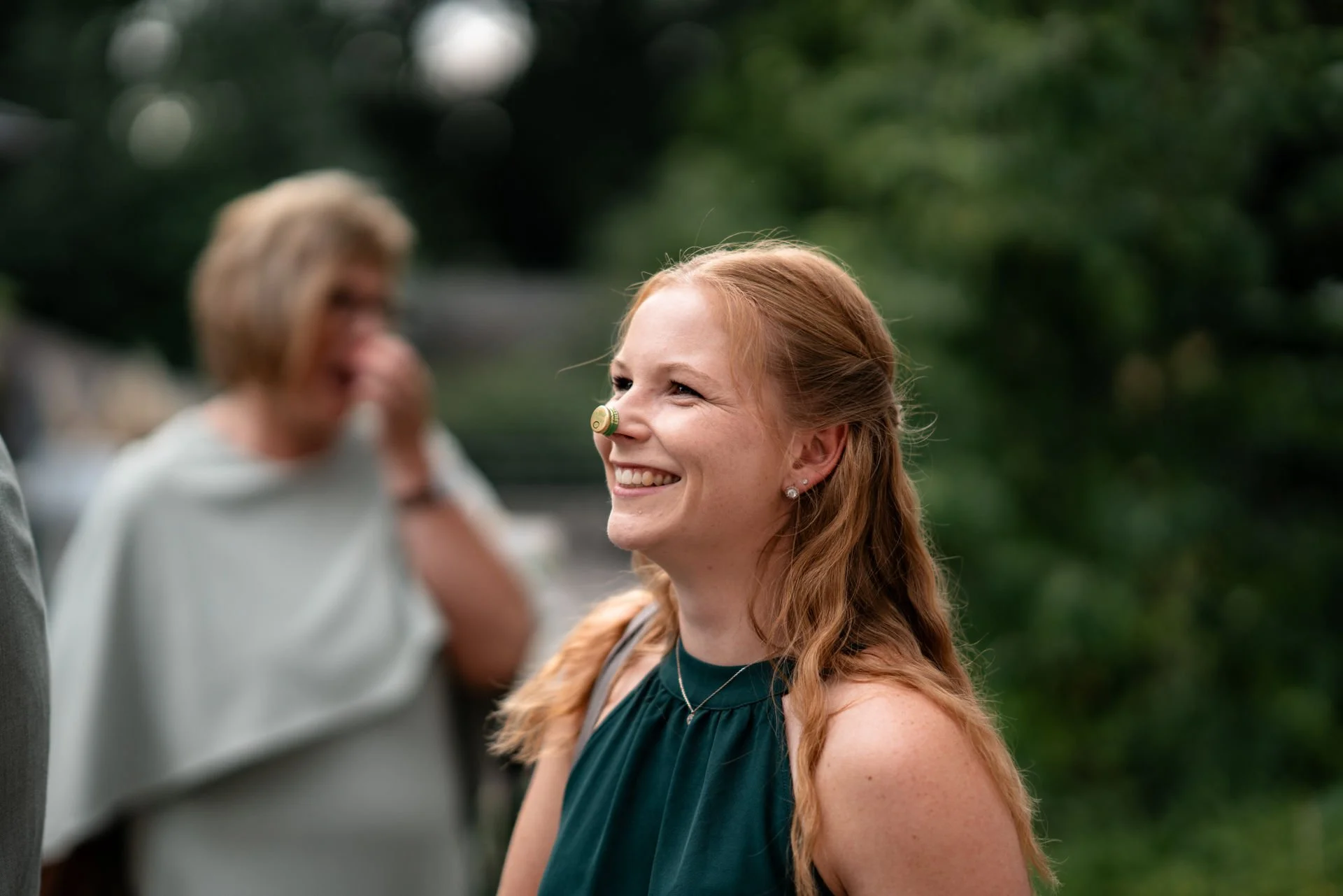 Lächelnde junge Frau mit roten Haaren trägt eine kleine Flasche auf der Nase, im Hintergrund eine ältere Frau, die an einer Party im Freien teilnimmt.