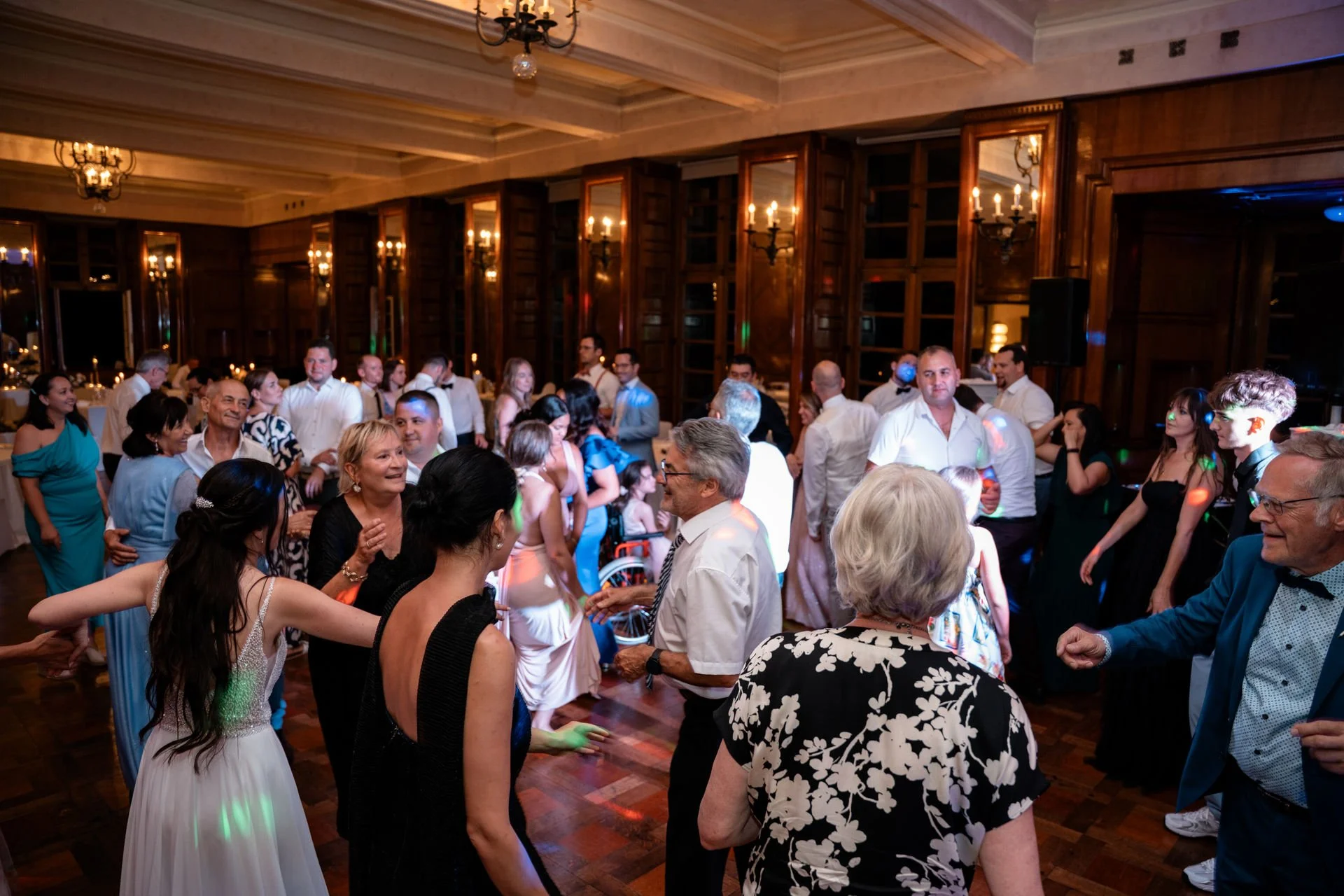Menschen tanzen und feiern in einem eleganten festlichen Raum mit dunkler Holzvertäfelung, Kronleuchtern und Spiegeln an den Wänden.
