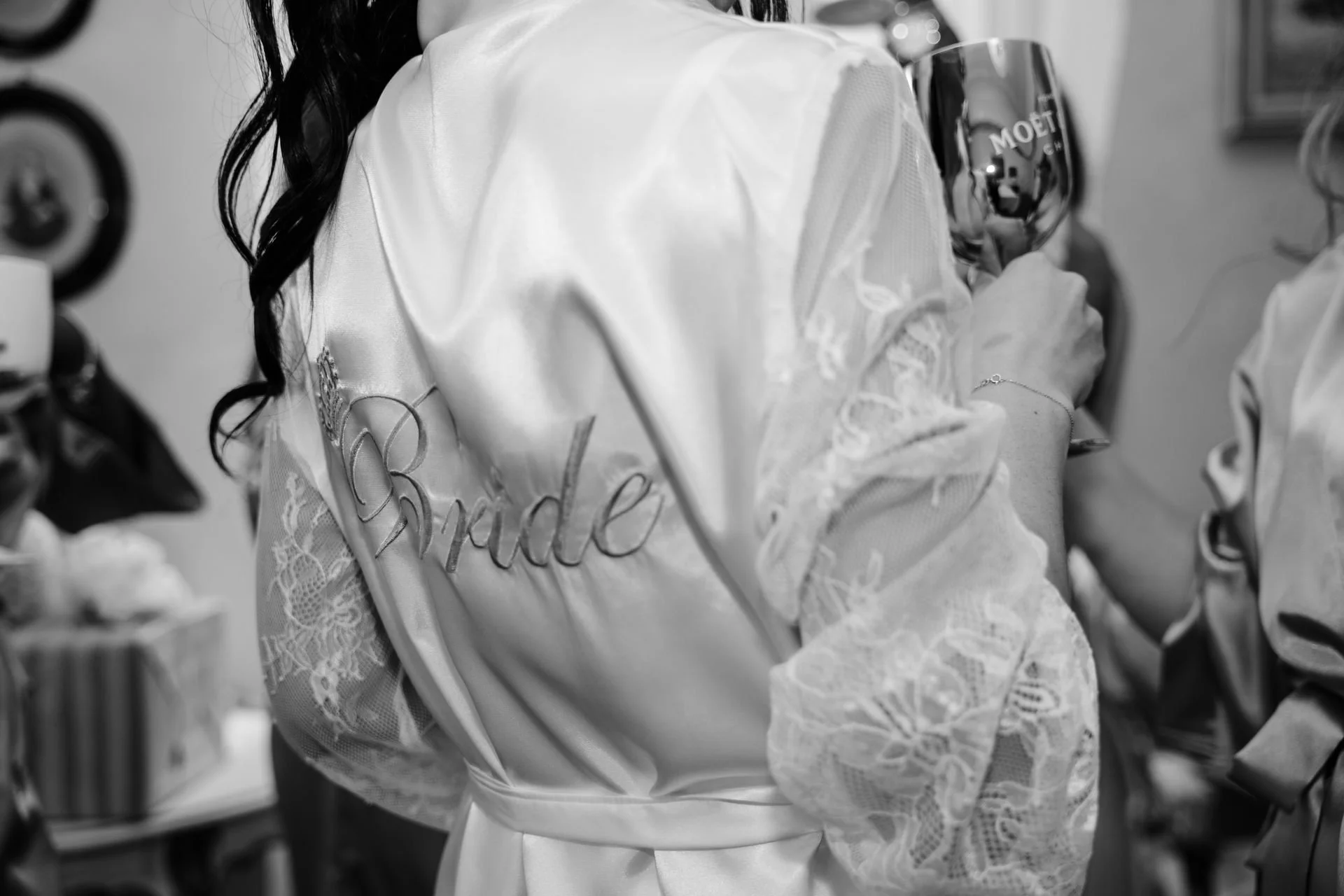 Nahaufnahme einer Frau in einem eleganten, satinähnlichen Bademantel mit dem Wort "Bride" (Braut) auf der Rückseite, die ein Weinglas hält.