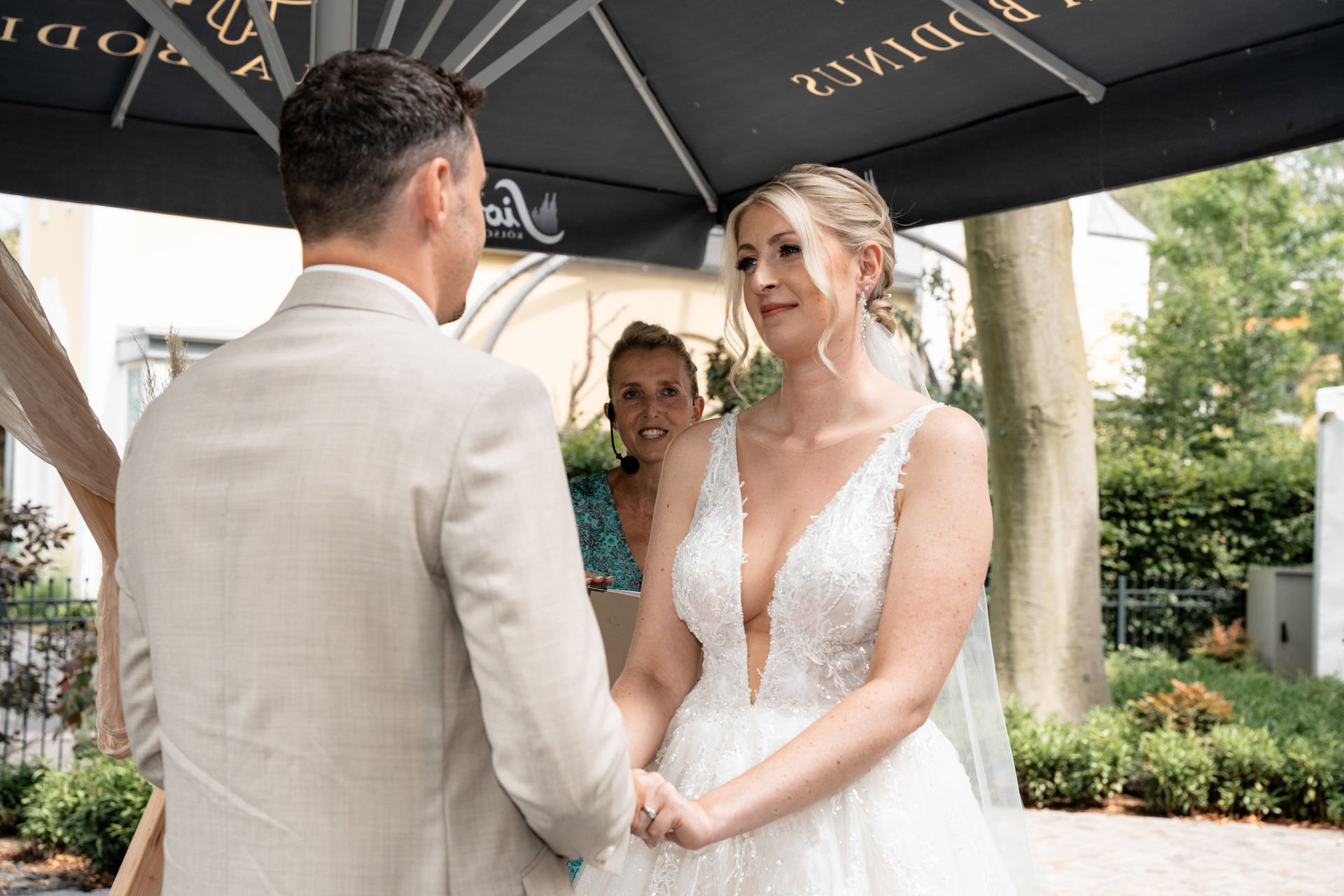 Braut und Bräutigam bei ihrer standesamtlichen Heirat unter einem schwarzen Sonnenschirm im Freien, mit standesamtlicher Zeremonie. Sie halten sich an den Händen, die Braut trägt ein weißes Hochzeitskleid mit tiefem V-Ausschnitt, der Bräutigam trägt 
