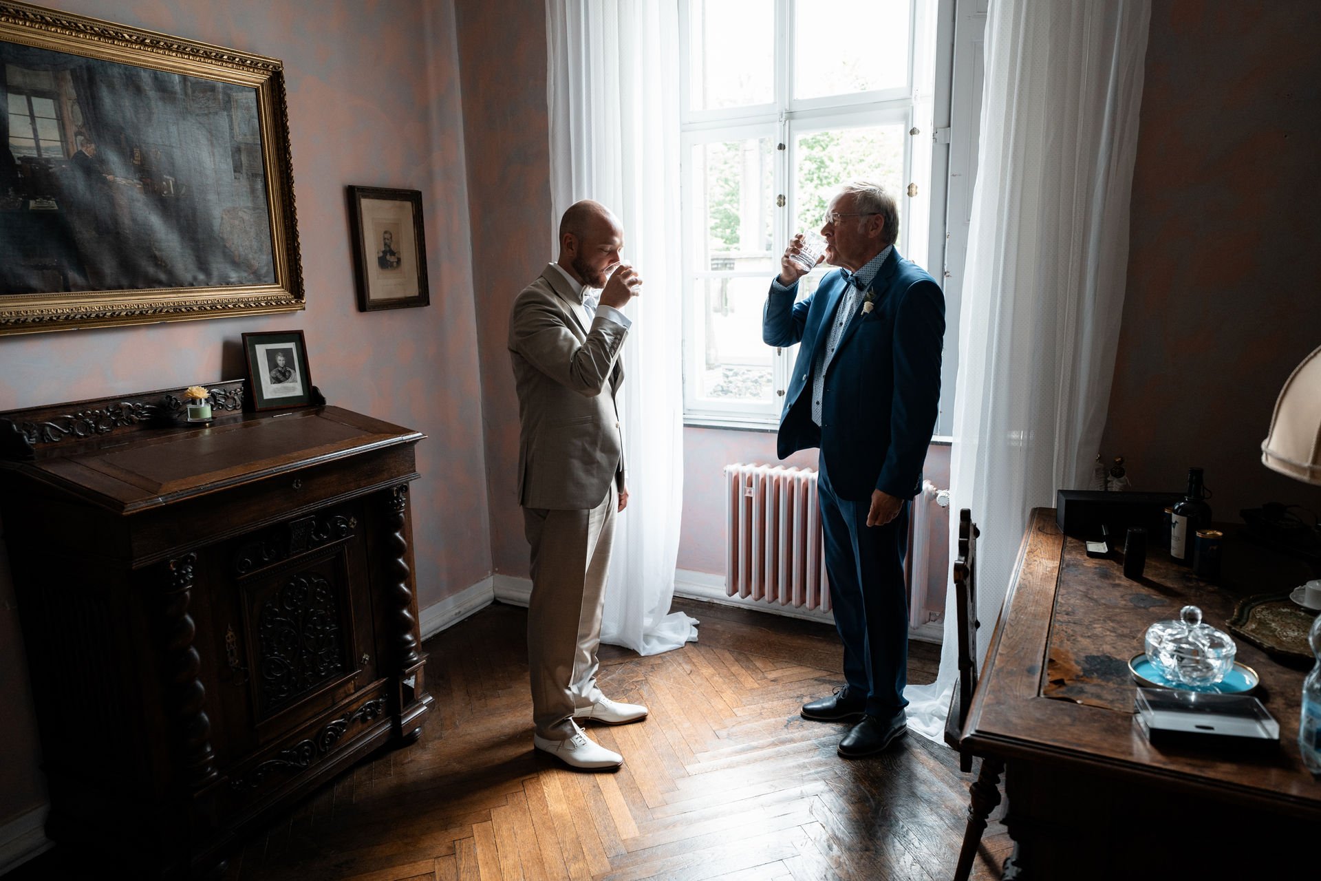 Zwei Männer in Anzügen trinken in einem hellen Raum, der mit klassischen Möbeln und Bildern geschmückt ist.