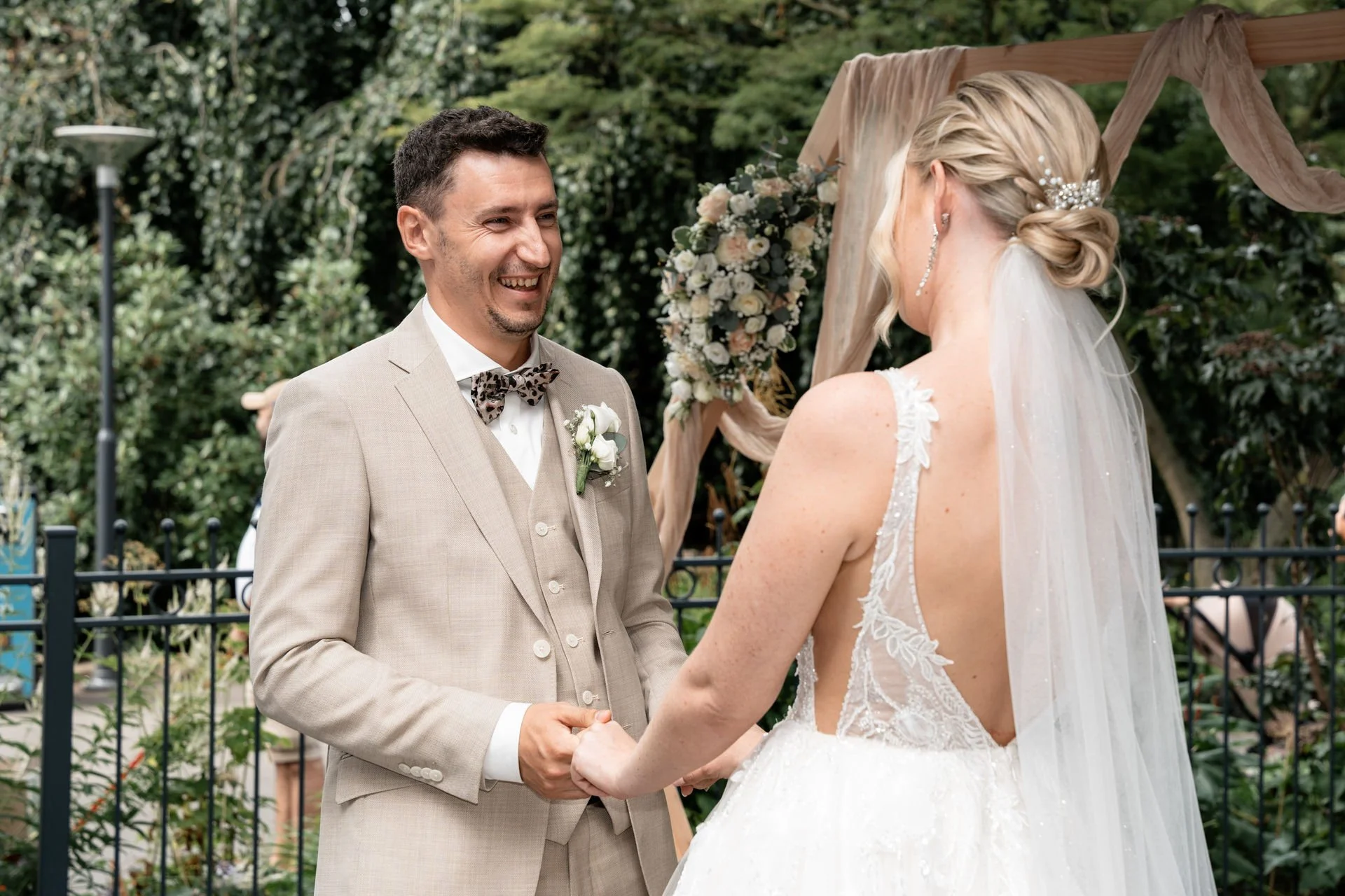 Ein Brautpaar beim Ja-Wort, die Braut trägt ein trägerloses, weißen Hochzeitskleid mit Spitze und Schleier, der Bräutigam trägt einen beige Anzug mit Fliege.