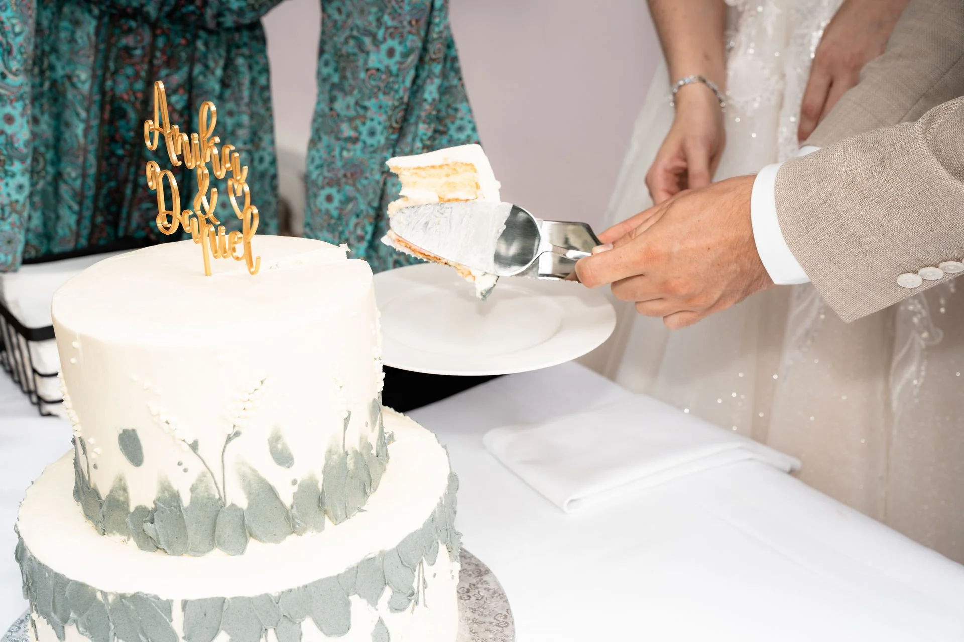 Ein Mann schneidet eine Hochzeitstorte, die mit weißen und grauen Dekorationen verziert ist, während andere Personen im Hintergrund stehen.