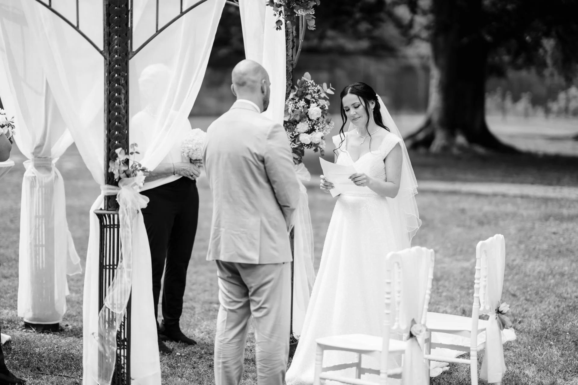 Eine Frau in einem weißen Brautkleid liest bei einer Hochzeit im Freien vor einem Mann in einem Anzug. Im Hintergrund sind weitere Personen und ein Baum zu sehen, die zeremoniell geschmückt sind.