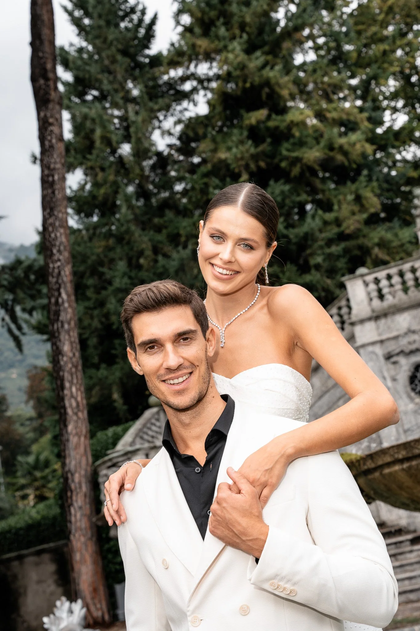 Ein glückliches Paar bei einer Hochzeit im Freien, mit grünen Bäumen im Hintergrund und einem historischen Gebäude oder Brunnen. Am Comer See