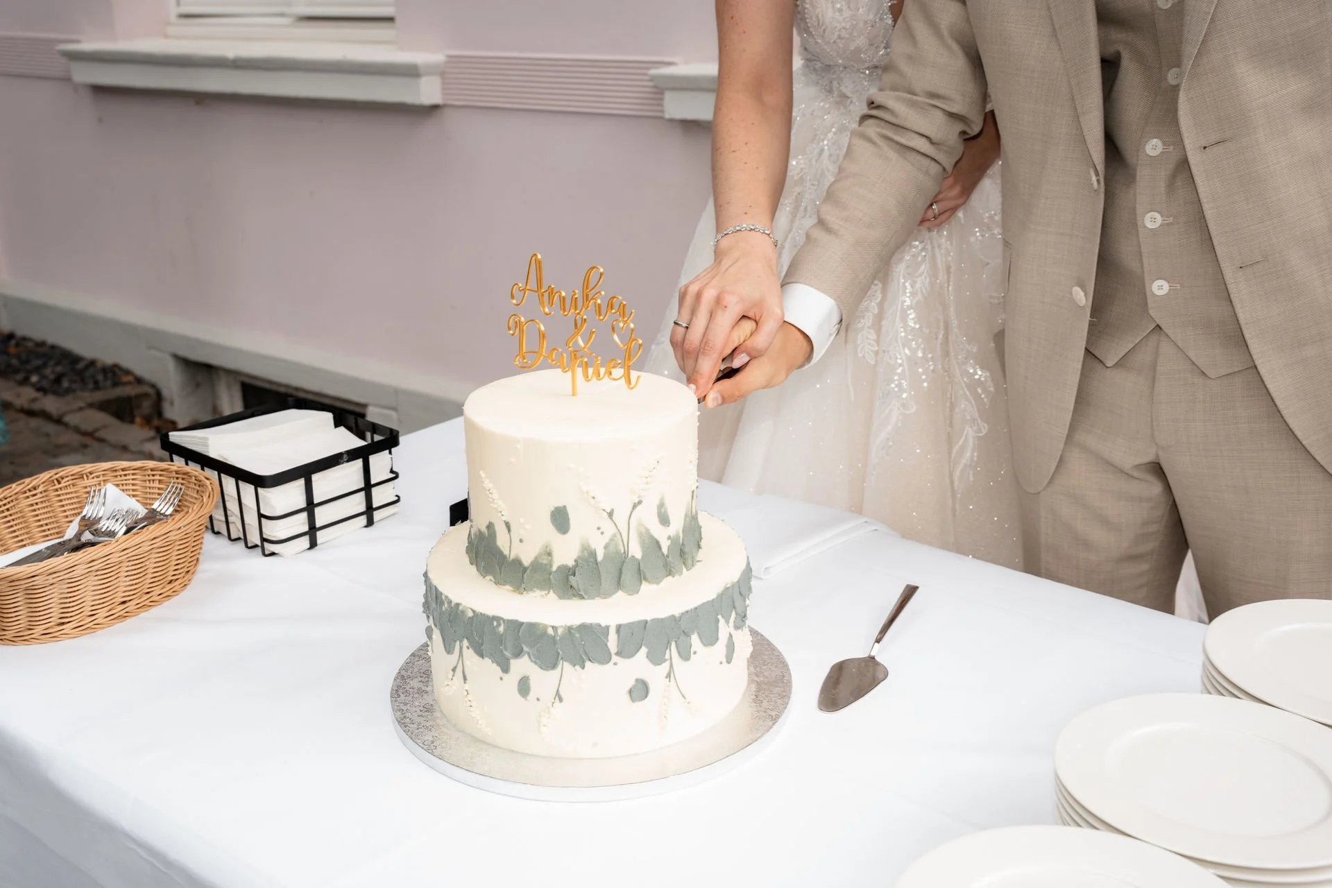 Ein Brautpaar schneidet eine Hochzeitstorte in Weiß mit grauen Akzenten. Auf der Torte befindet sich eine goldene Dekoration mit den Namen 'Anuja & Daniel'.