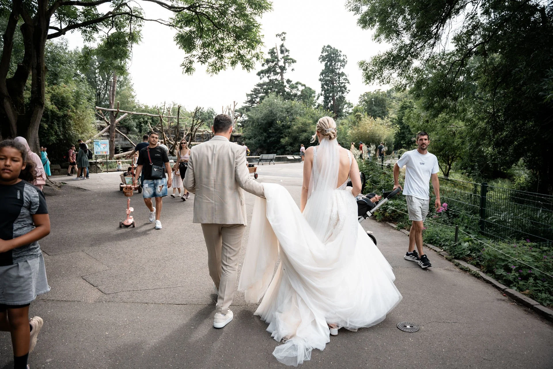 Braut und Bräutigam gehen in einem Park spazieren, umgeben von anderen Besuchern und Bäumen.