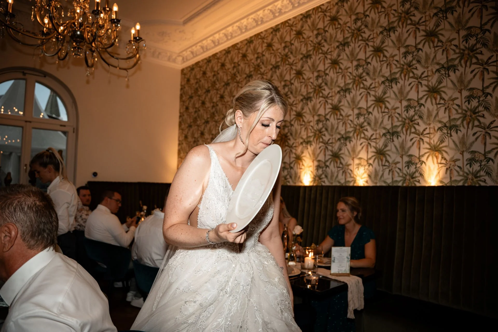 Eine Hochzeitsgesellschaft, bei der eine Braut in einem weißen Kleid auf einem Festtisch steht und ihr Brautkleid auf ein Tablett legt, während um sie herum Gäste sitzen.