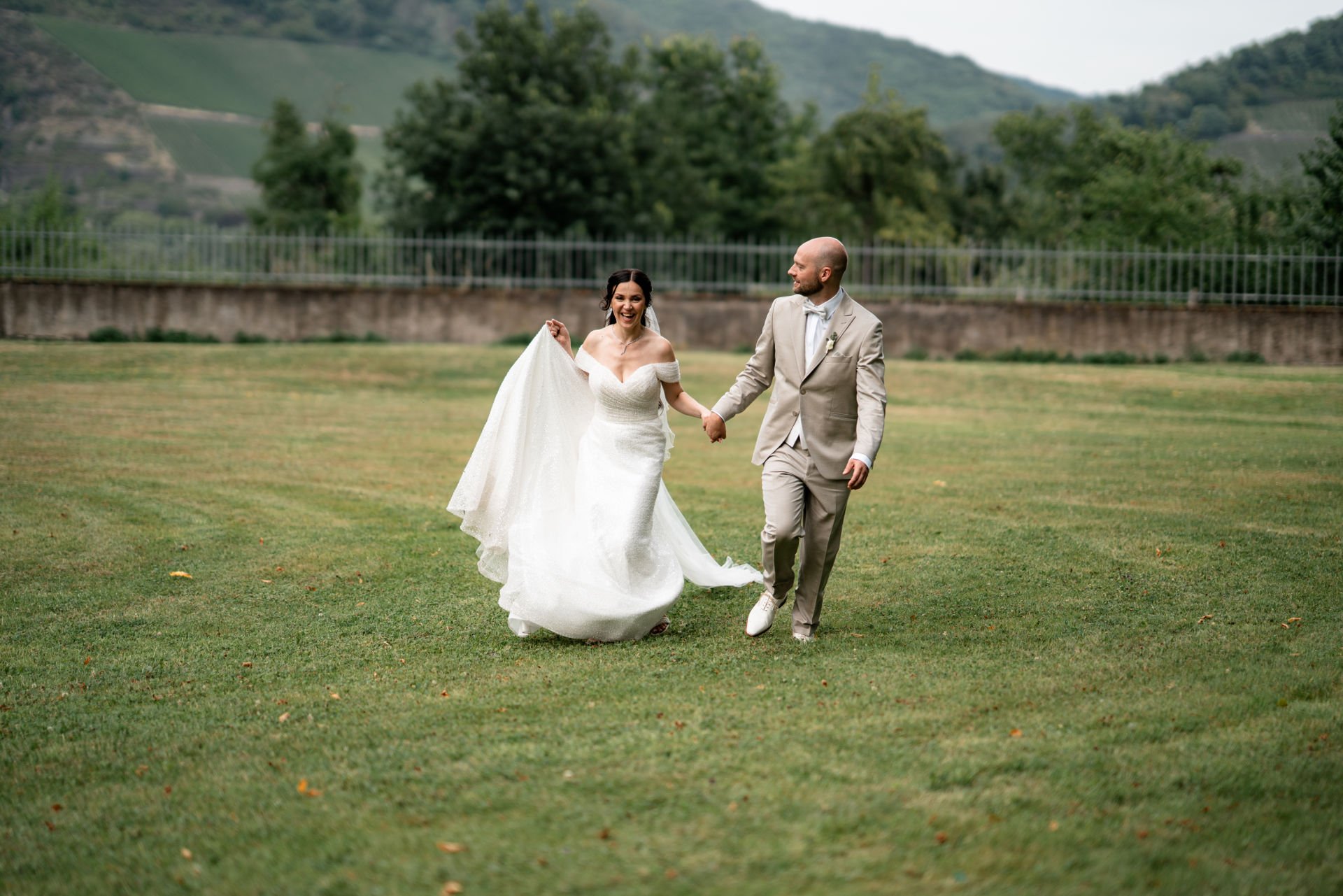 Ein lächelndes Brautpaar beim Spaziergang auf einem grünen Feld, im Hintergrund Bäume und Hügel.