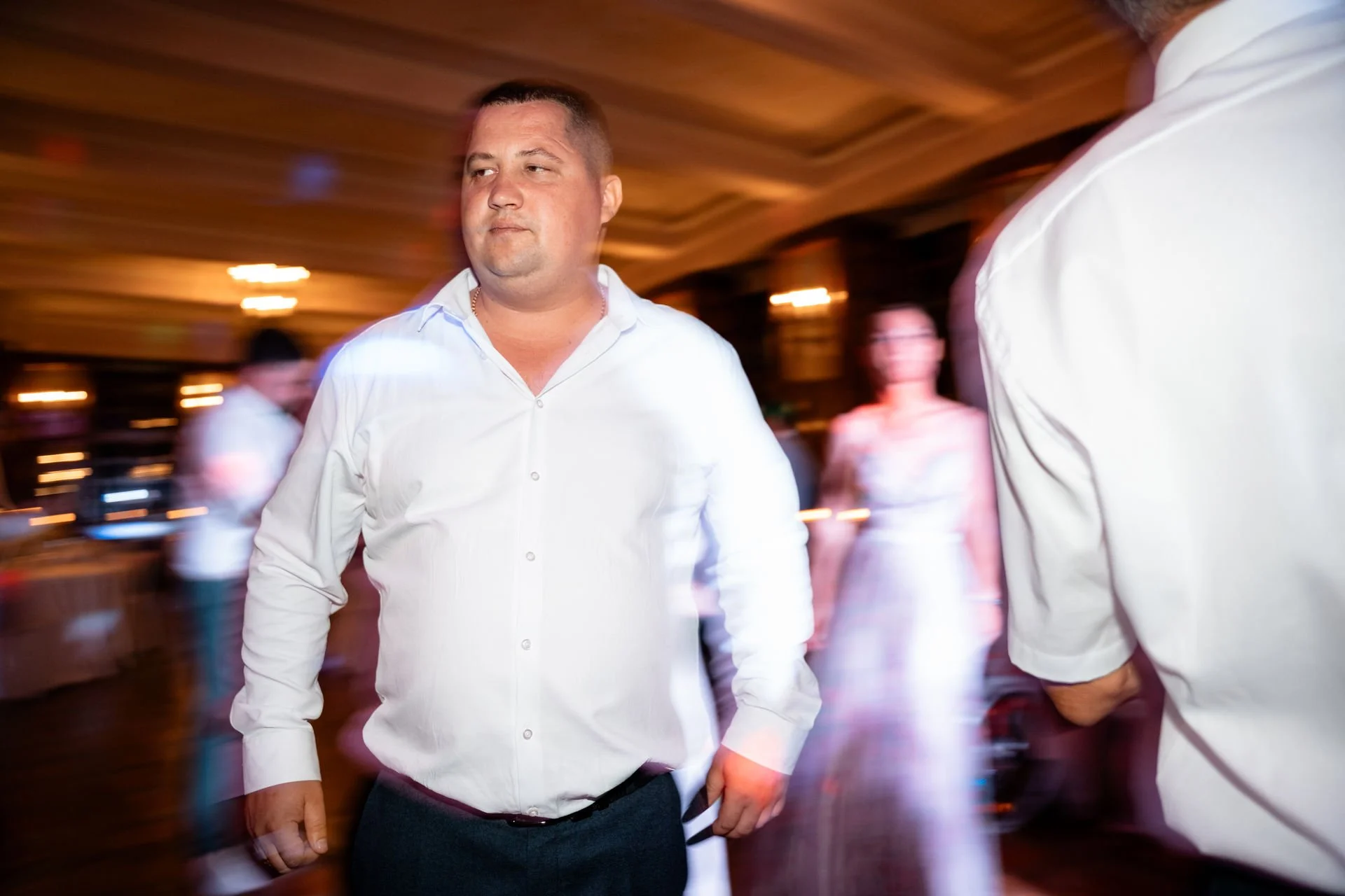 Mann in Weißes Hemd bei Tanzveranstaltung, hinter ihm bunte Lichter und weitere tanzende Personen, Bewegung und Atmosphäre eines Balls oder Festes.