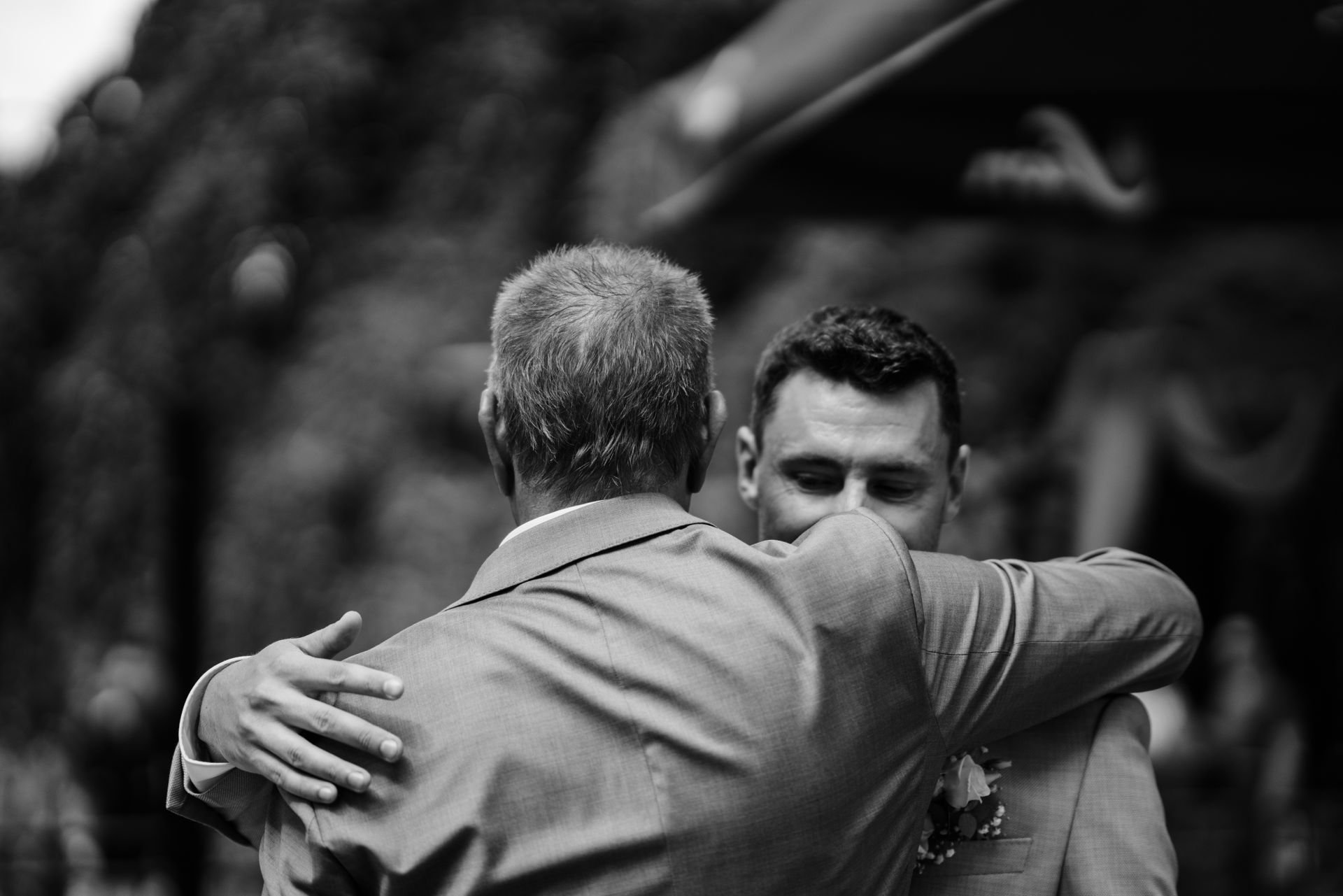 Zwei Männer umarmen sich bei einer Hochzeitsfeier, einer trägt einen Anzug, der andere den Hochzeitsanzug, in schwarz-weiß aufgenommen.