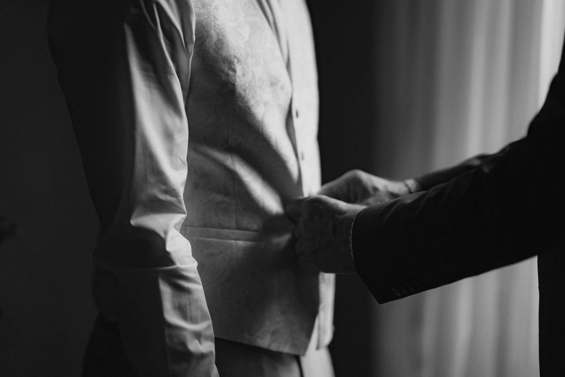 Schwarz-weiß-Bild zeigt zwei Personen, das eine fixiert eine Anzugjacke und das andere hilft beim Zuknöpfen.