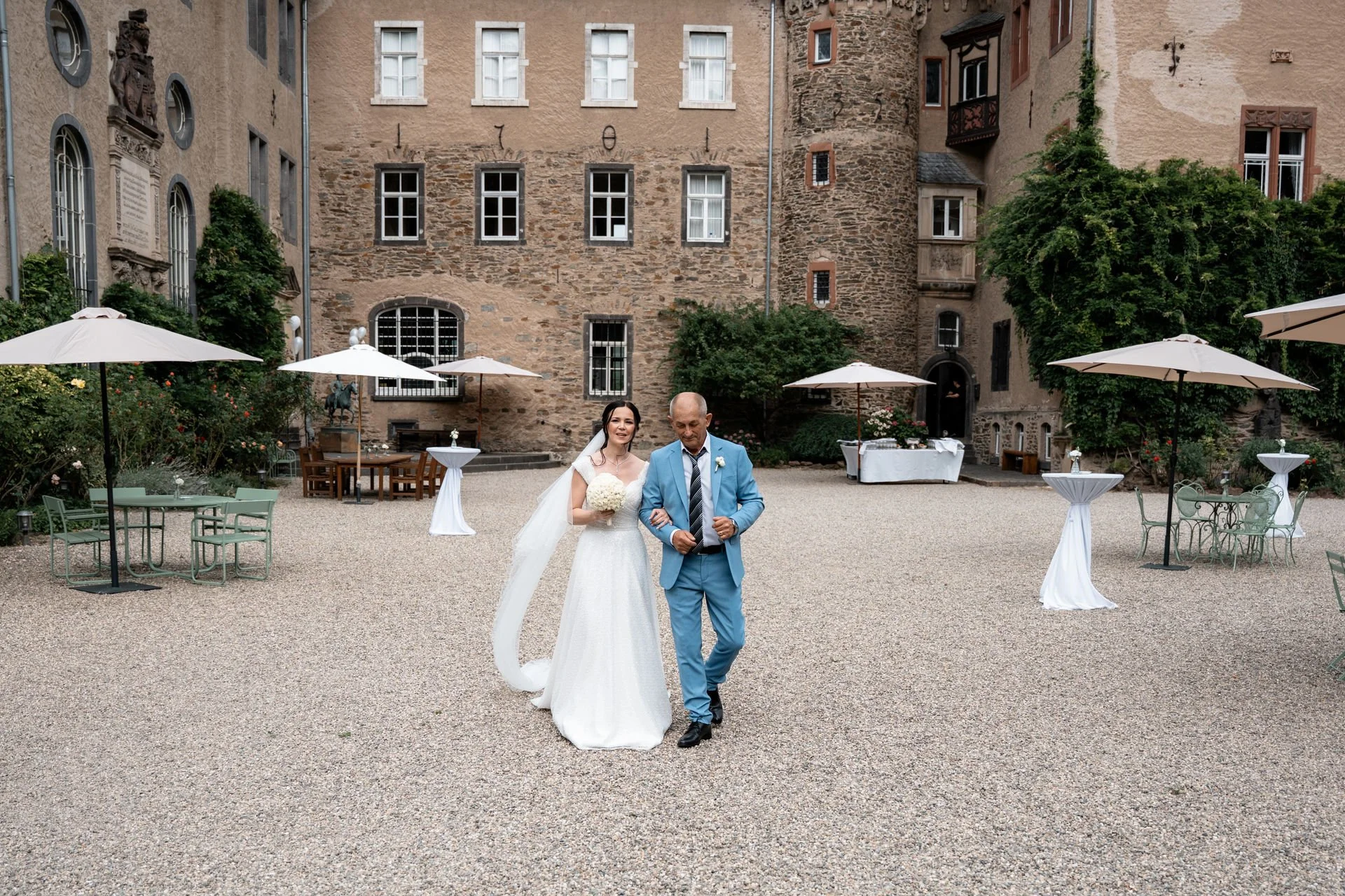 Braut in weißem Hochzeitskleid mit Blumenstrauß und ihr Vater in einem blauen Anzug gehen auf einem gepflasterten Hof vor einem alten Stein- und Fachwerkhaus.