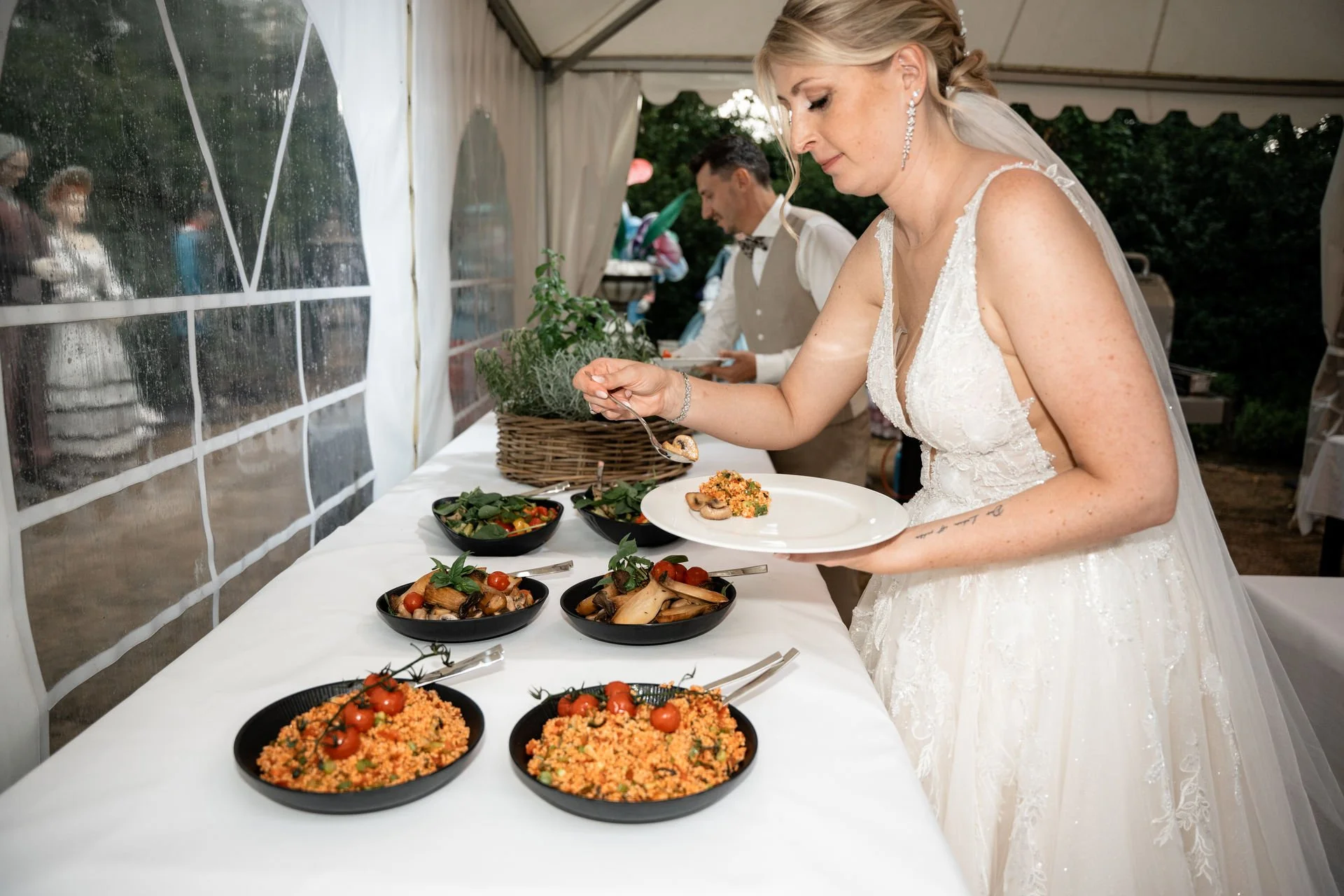 Braut in weißen Hochzeitskleid beim Buffet, serviert sich Salate und Beilagen, im Zelt bei einer Feier, mit anderen Gästen im Hintergrund.