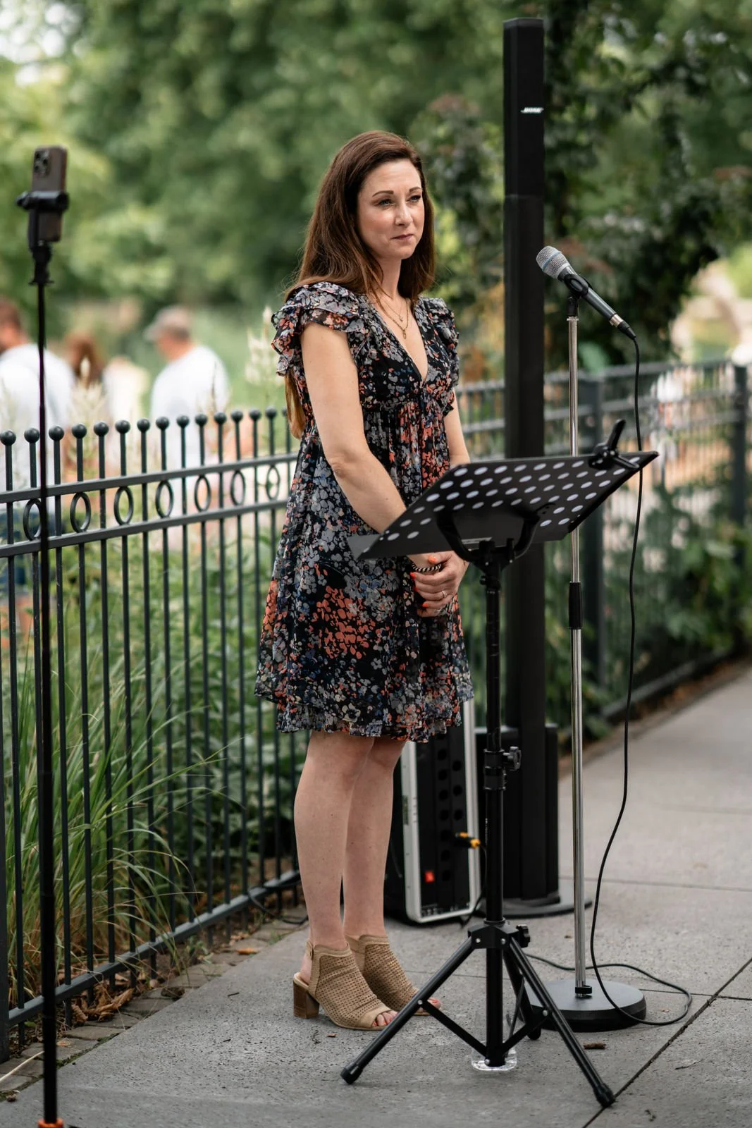 Eine Frau steht neben einem Mikrofon und einem Notenständer, während sie bei einem Outdoor-Event spricht oder singt.