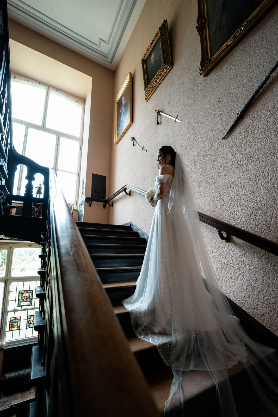 Braut in Hochzeitskleid steht auf der Treppe im Profil, hält einen Blumenstrauß, vor einer Wand mit Gemälden und • einer großen Fensterfront, durch die Tageslicht fällt.