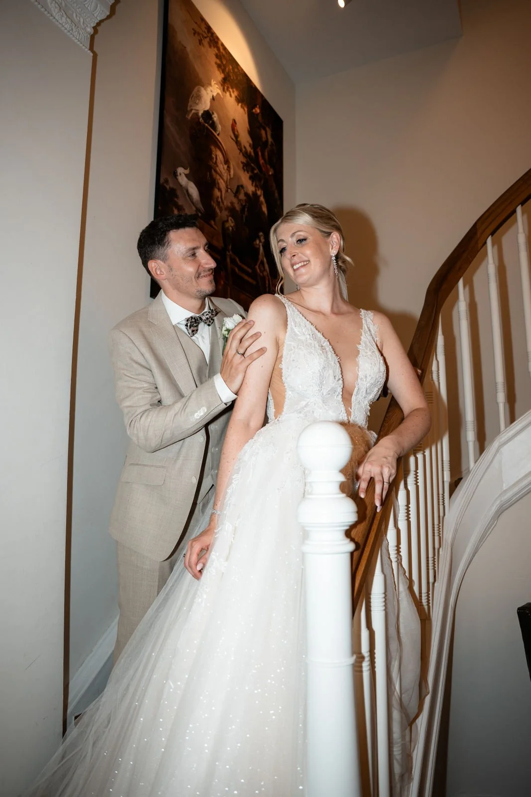 Ein Brautpaar steht auf einer Treppe, die Braut trägt ein weißes, elegante Brautkleid mit tiefem Ausschnitt, der Bräutigam trägt einen hellen Anzug mit Fliege. Sie lachen und schauen sich liebevoll an.