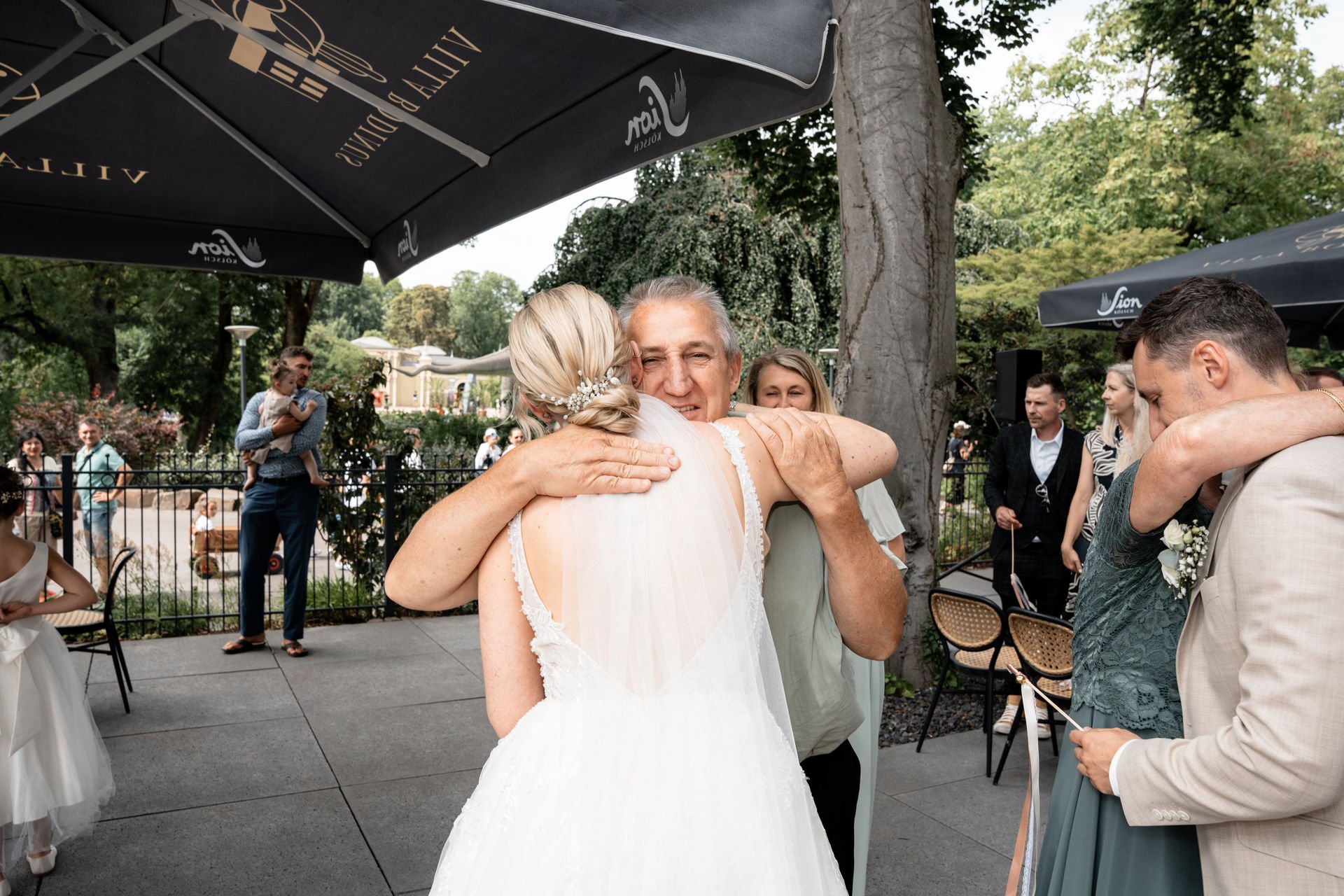 Ein Hochzeitsbild mit einem Brautpaar, das sich herzlich umarmt, umgeben von Gästen im Freien. Im Hintergrund sind Bäume und Sonnenschirme sichtbar.