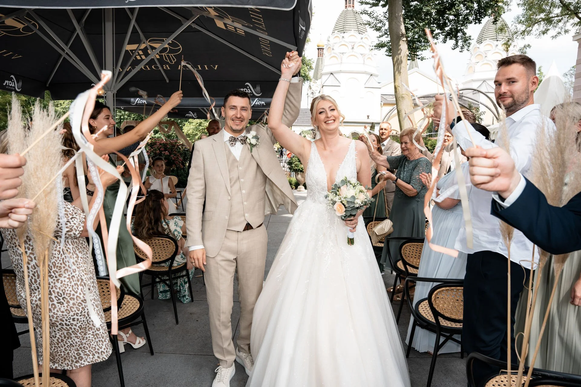 Braut und Bräutigam feiern ihre Hochzeit im Freien, umgeben von Freunden und Familie, die mit bunten Bändern winken, während die beiden glücklich lachen.