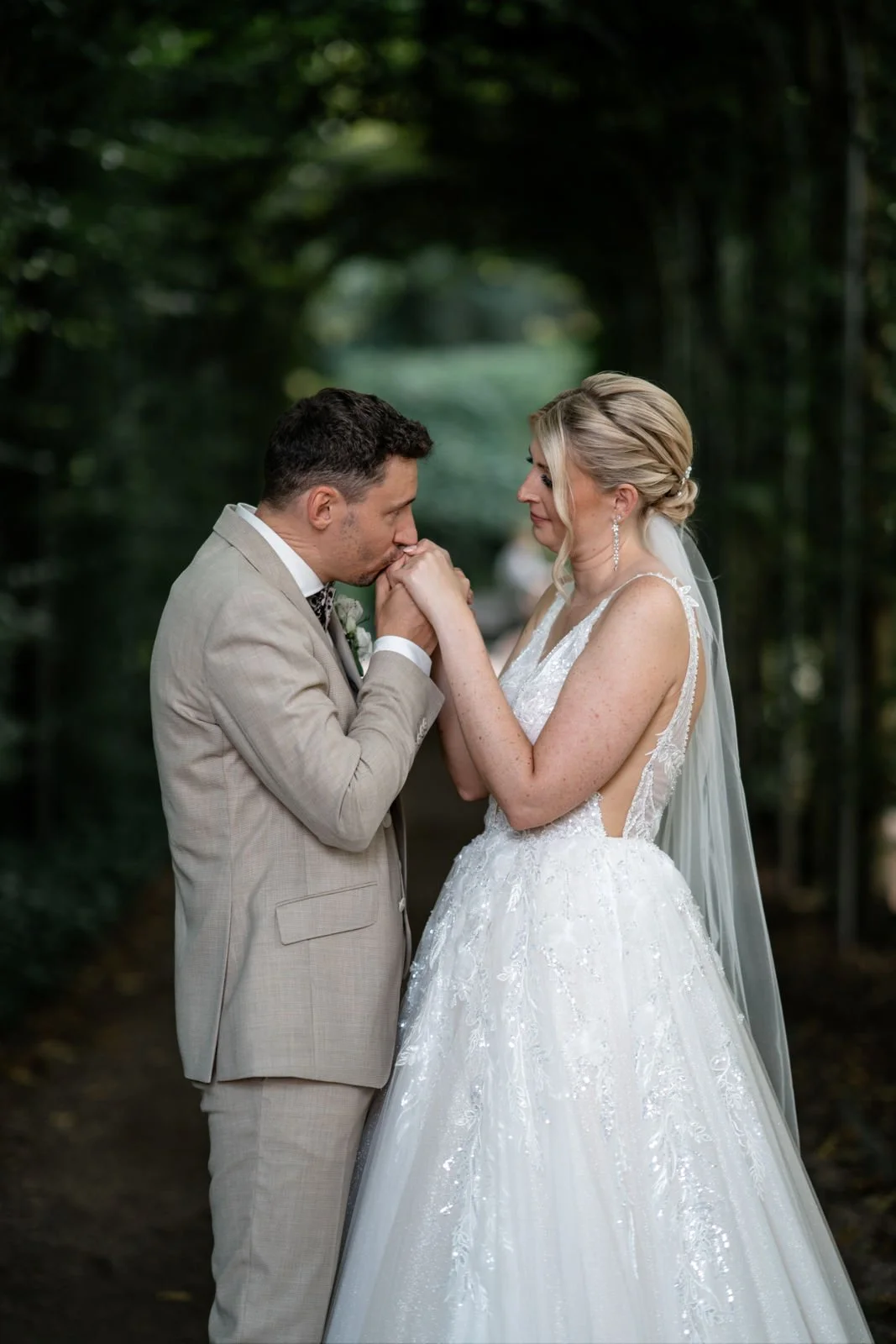 Ein Brautpaar in einem Wald, der Bräutigam küsst die Hand der Braut, beide tragen Hochzeitskleidung.