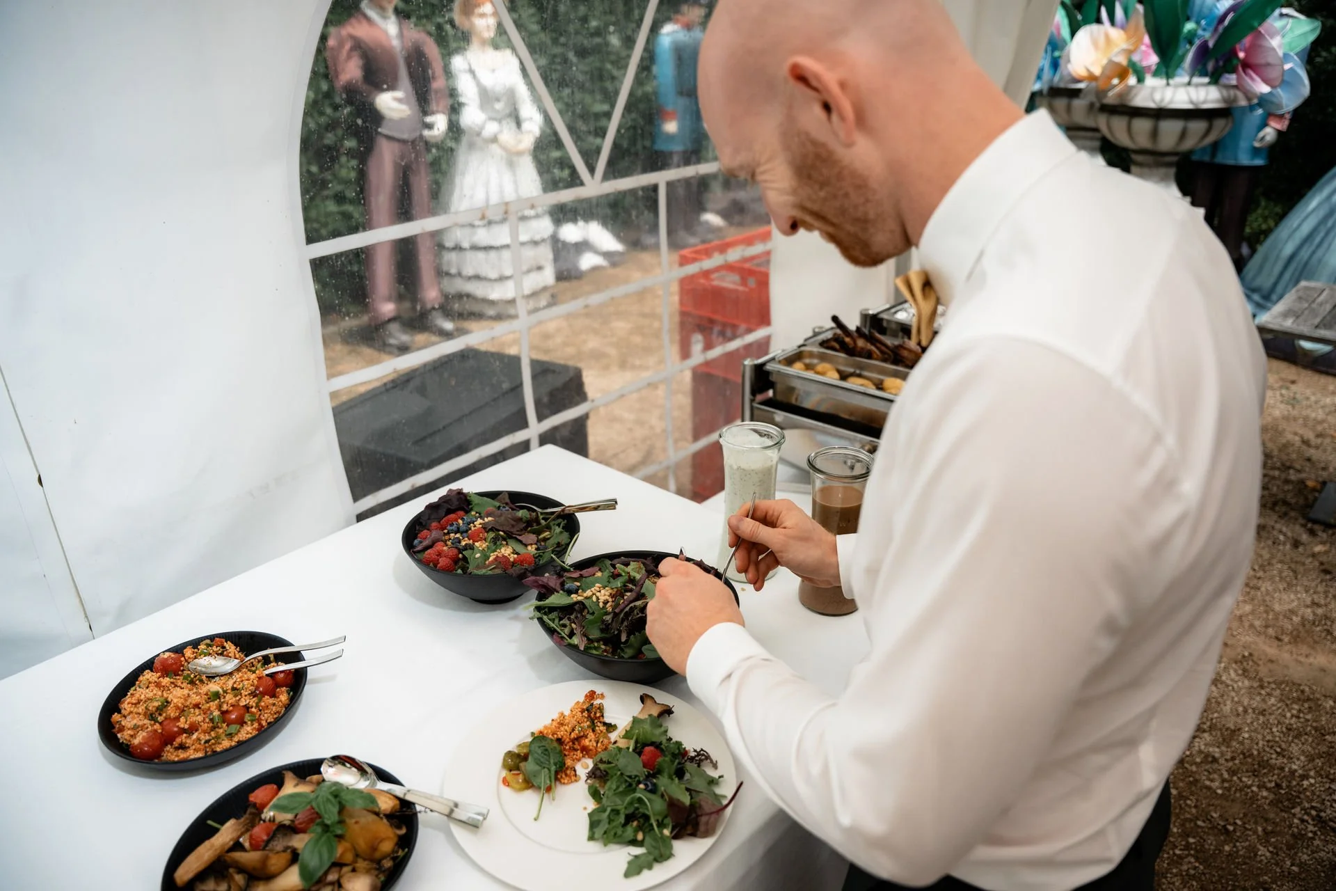 Ein Mann in weißem Hemd bereitet Salate an einem Buffet bei einer Hochzeit vor, im Hintergrund sind zwei Hochzeitsfiguren im Freien sichtbar.