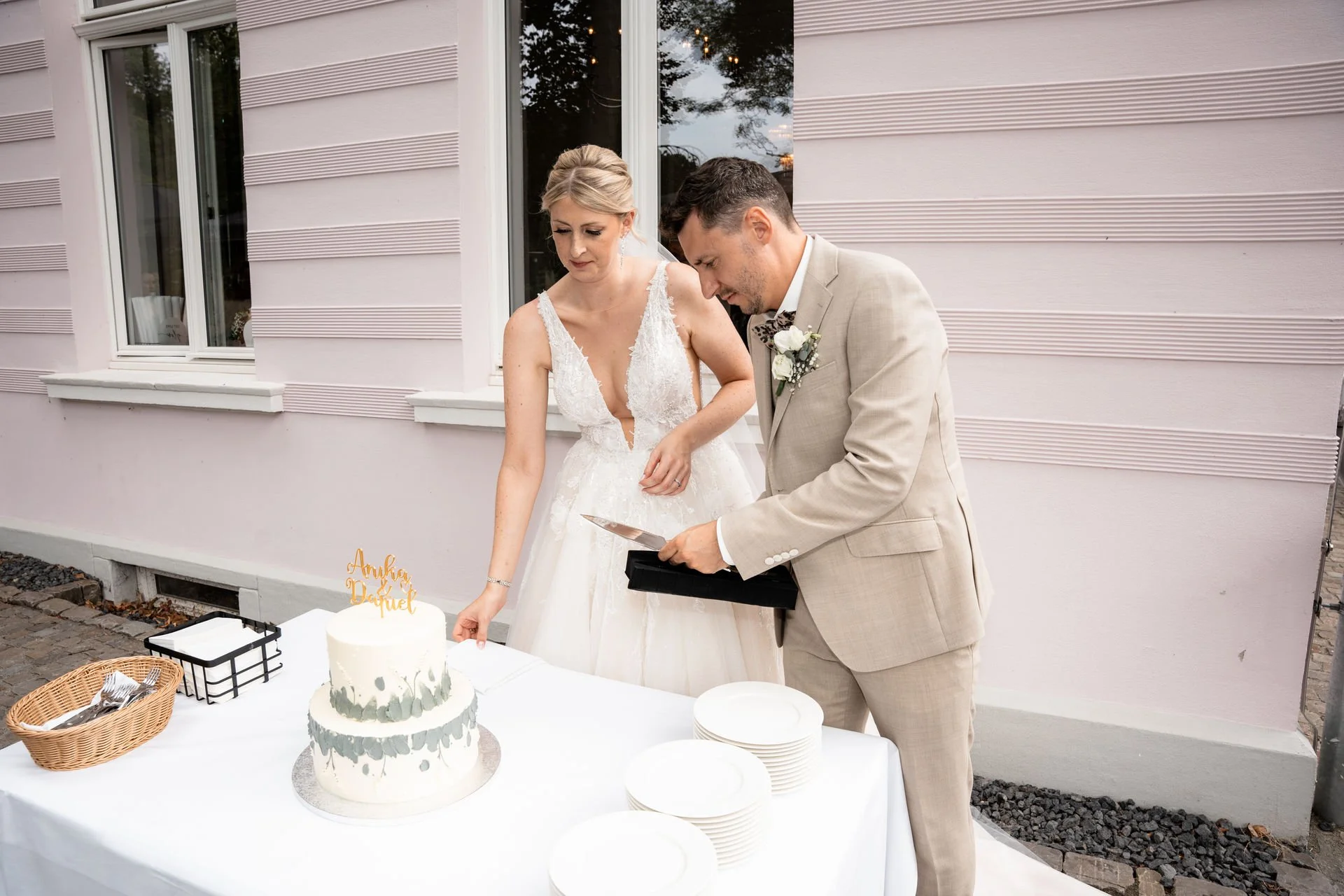 Ein Brautpaar schneidet eine Hochzeitstorte gemeinsam an, das Brautkleid ist weiß und das Brautpaar trägt elegante Hochzeitskleidung, draußen vor einem Haus