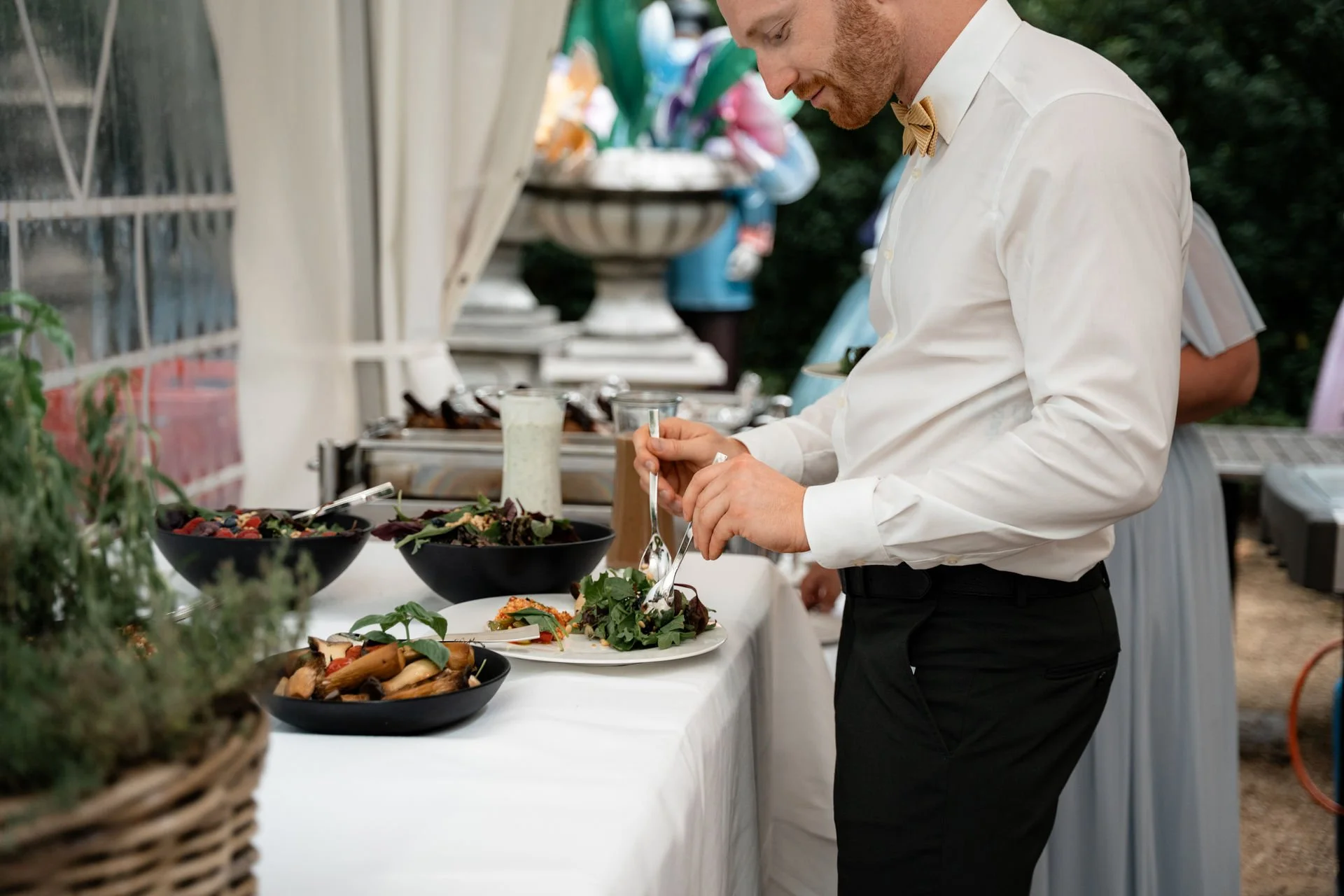 Ein Mann in Weiß mit Fliege bedient sich an einem Buffettisch mit Salaten und anderen Speisen im Freien.