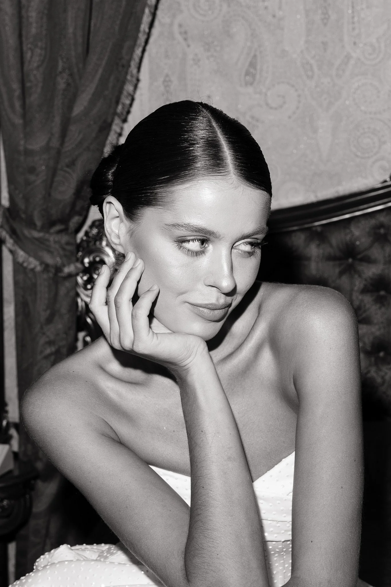 Schwarz-weiß-Fotografie eines jungen Frauenporträts, die elegante Frisur und Make-up trägt, mit einem handabdruck auf der Wange, in einem stilvollen Raum.