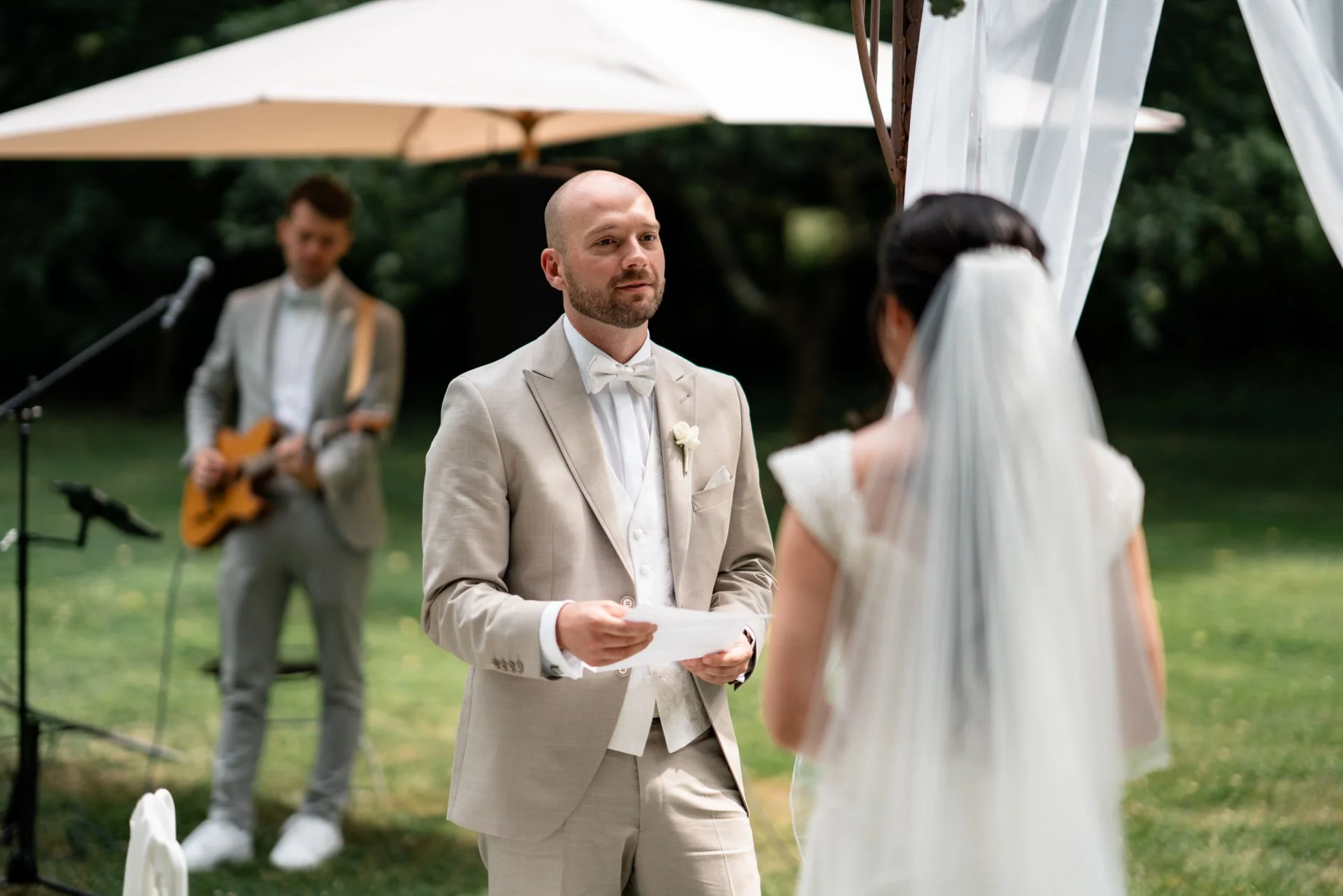 Ein Brautpaar bei einer Hochzeitszeremonie im Freien, mit einem Mann, der das Gelübde liest, und einer Frau mit Schleier, die ihm gegenübersitzt, im Hintergrund ein Musiker mit Gitarre.