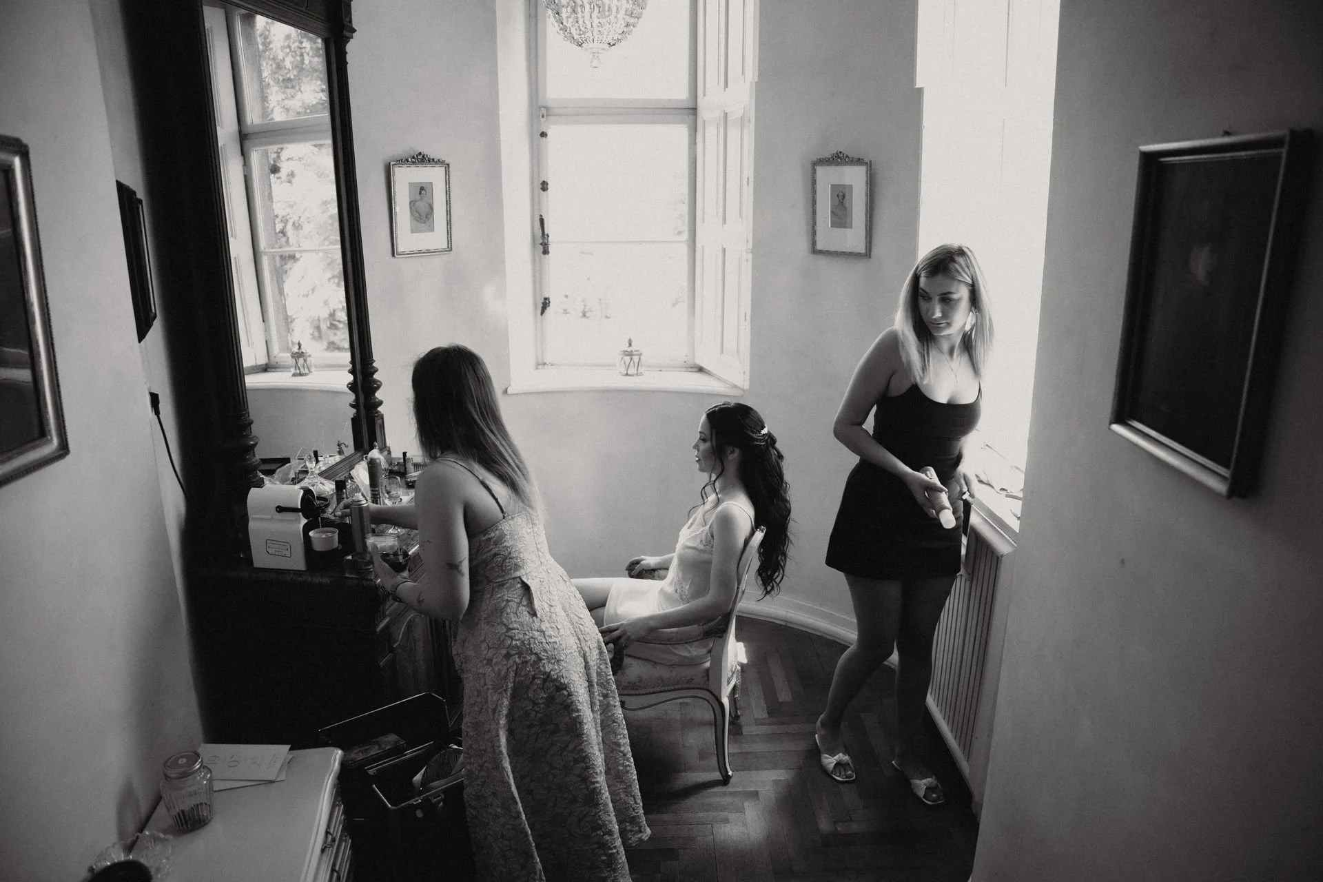 Drei Frauen bereiten sich in einem hellen Raum vor, während eine frischt sich bei einem Schminktisch auf, eine sitzt mit langen schwarzen Haaren und weißen Kleid, die dritte steht an einem Fenster, die eine Frau hilft, sich fertig zu machen.