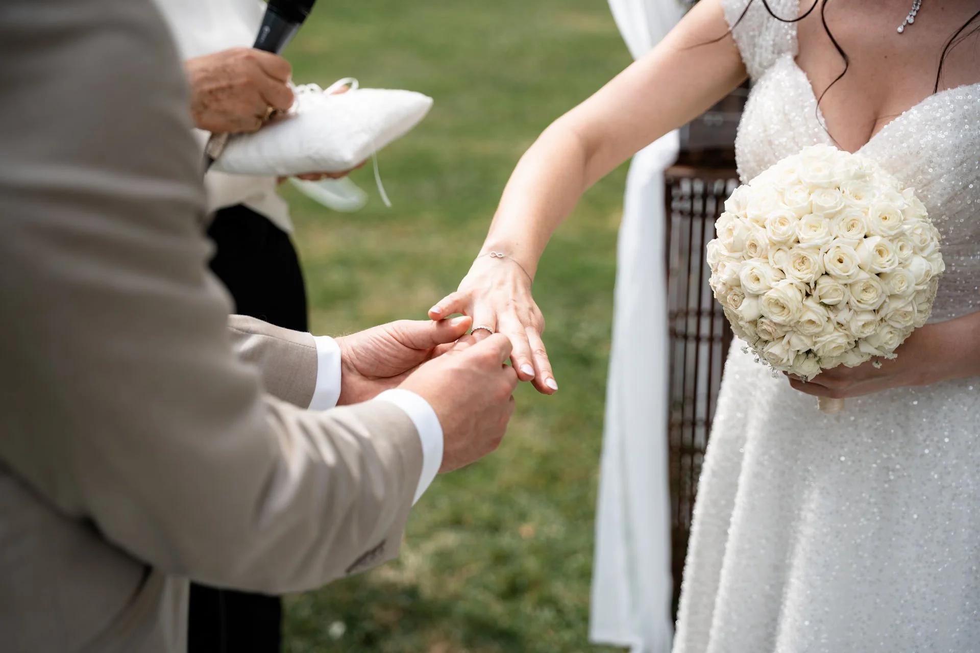 Ein Brautpaar bei einer Trauungszeremonie, der Bräutigam legt den Ehering an die Hand der Braut, die eine weiße Blumenvase hält und eine weiße Hochzeitstracht trägt, während ein Offizieller die Zeremonie leitet.