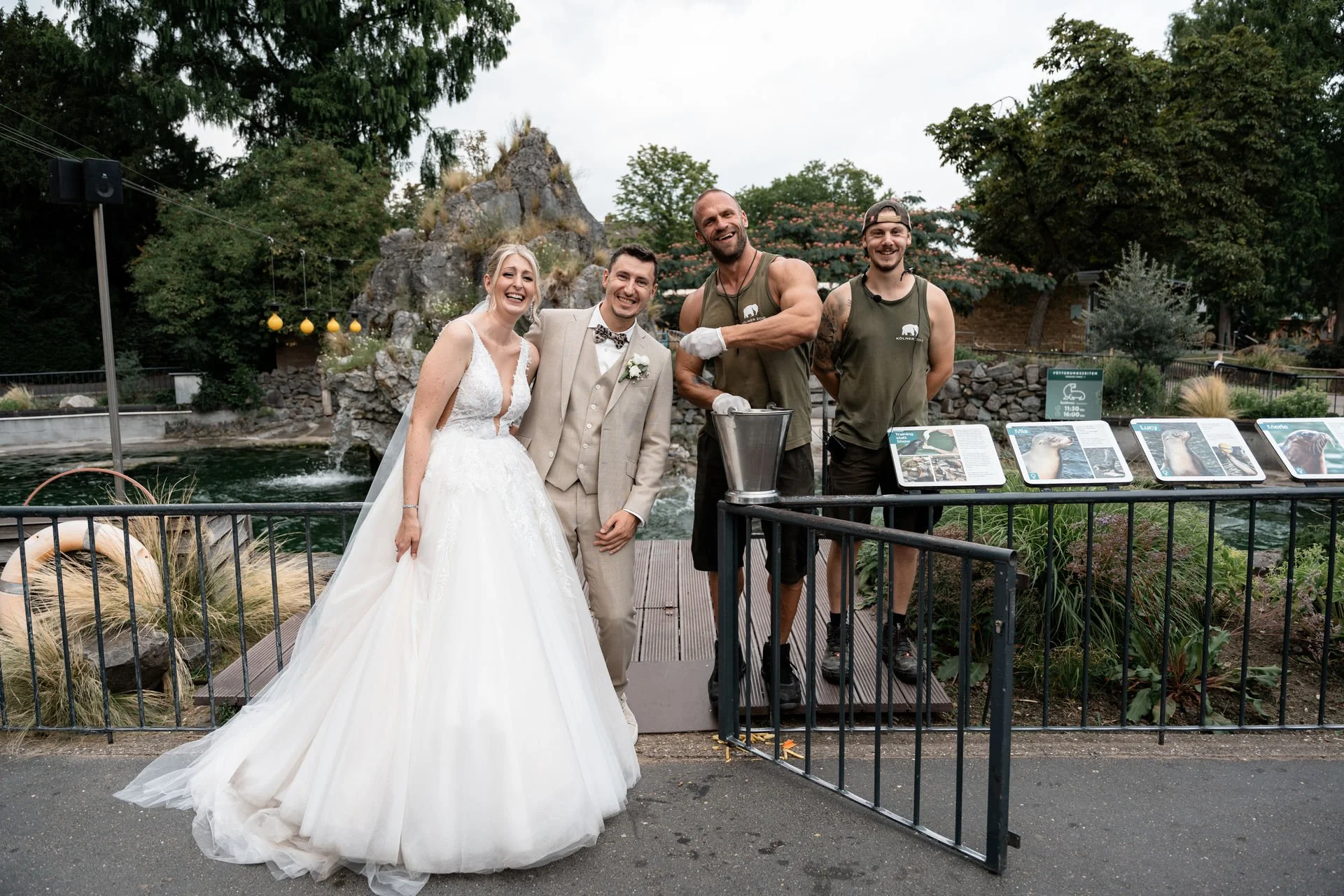 Hochzeitspaar mit zwei Arbeitern bei einer Tier-Attraktion im Freien, lachend und schmutzig, im Hintergrund Wasserfall und Bäume.