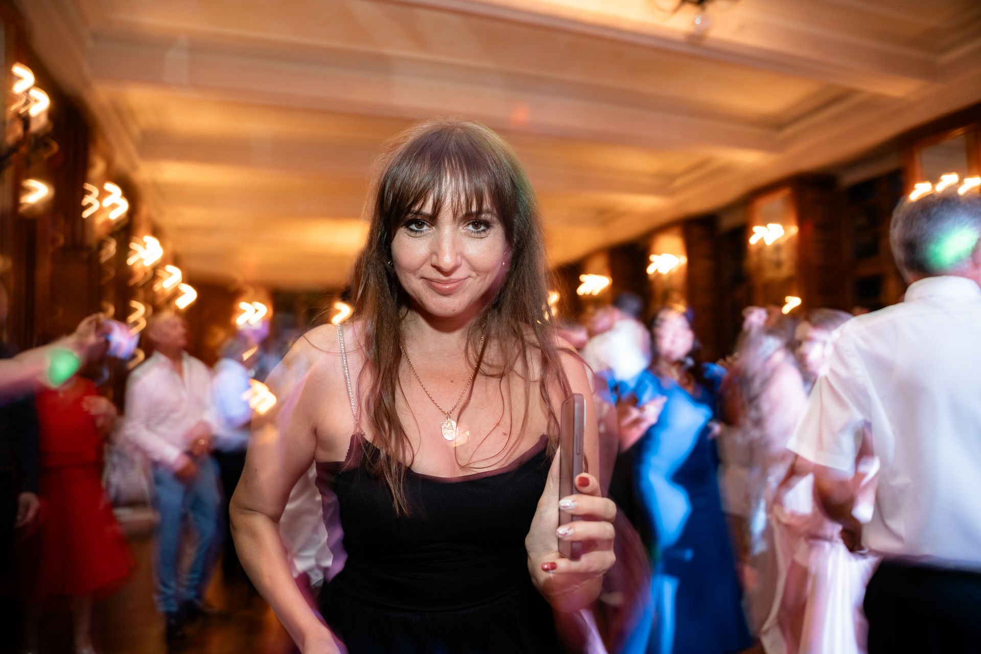 Eine Frau mit braunen Haaren und einem schwarzen Kleid, die auf einer Party tanzt, während sie ein Smartphone in der Hand hält. Im Hintergrund feiern mehrere Menschen in einem festlich geschmückten Raum.