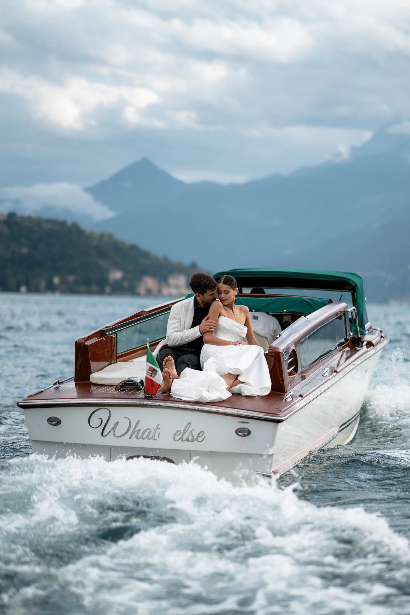 Paare sind auf einem Boot auf einem See, mit Bergen im Hintergrund, die Frau trägt ein weißes Kleid, der Mann trägt einen schwarzen Anzug, das Boot ist aus Holz gefertigt und hat eine kleine italienische Flagge.