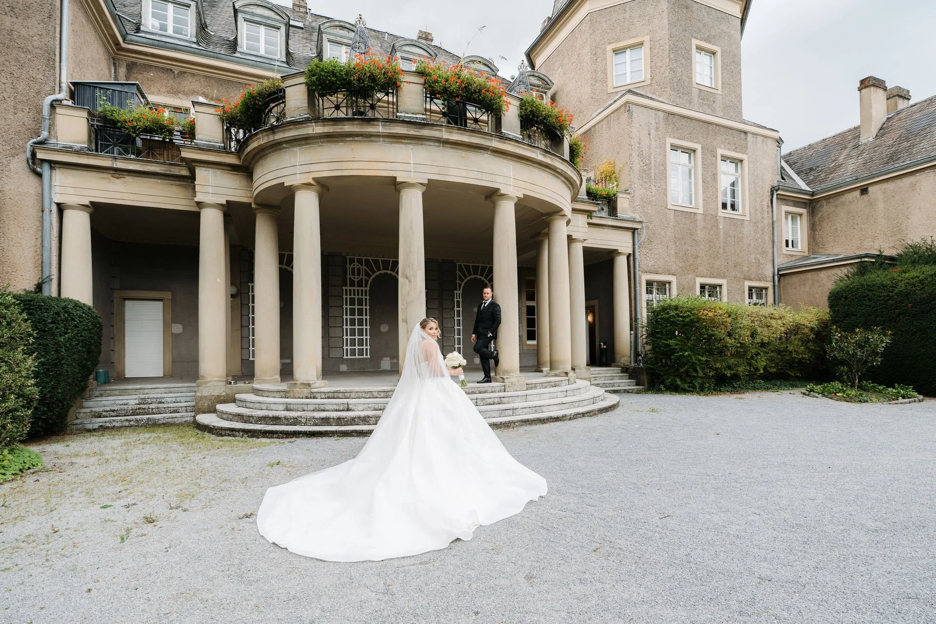 Ein Brautpaar posiert vor einem großen, historischen Haus mit einem verzierten Balkon, alten Säulen und einer Treppe. Die Braut trägt ein weißes Hochzeitkleid mit langem Schleier und hält einen Blumenstrauß. Der Bräutigam trägt einen schwarzen Anzug mit weißem Hemd und Krawatte.