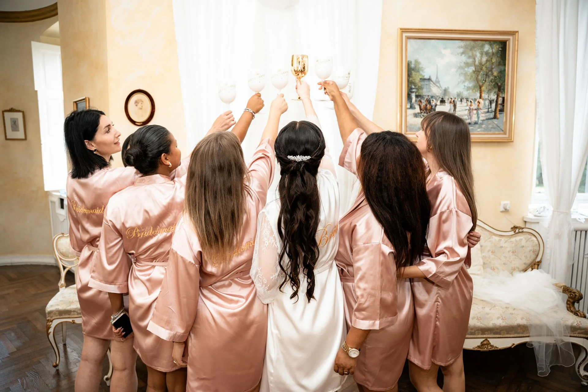 Eine Gruppe von Frauen, wahrscheinlich Brautjungfern und die Braut, heben mit Gläsern und einem Pokal an einem Hochzeitstag, in einem hellen Raum mit Kunstwerken an der Wand.