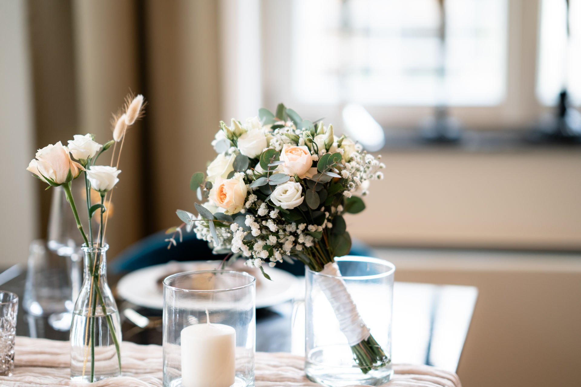 Blumenstrauß mit weißen und hellrosafarbenen Blumen auf einem Tisch, daneben Kerzen in Glasvasen.